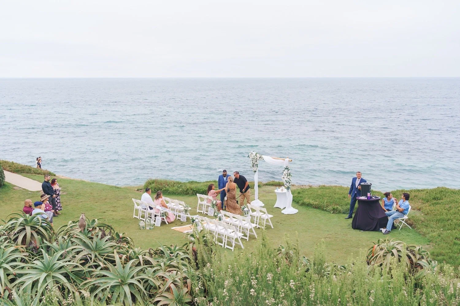 A wedding ceremony on a grassy lawn near the ocean, with guests seated and standing, a couple exchanging vows under an arch decorated with white fabric and flowers, with a DJ setup to the side.