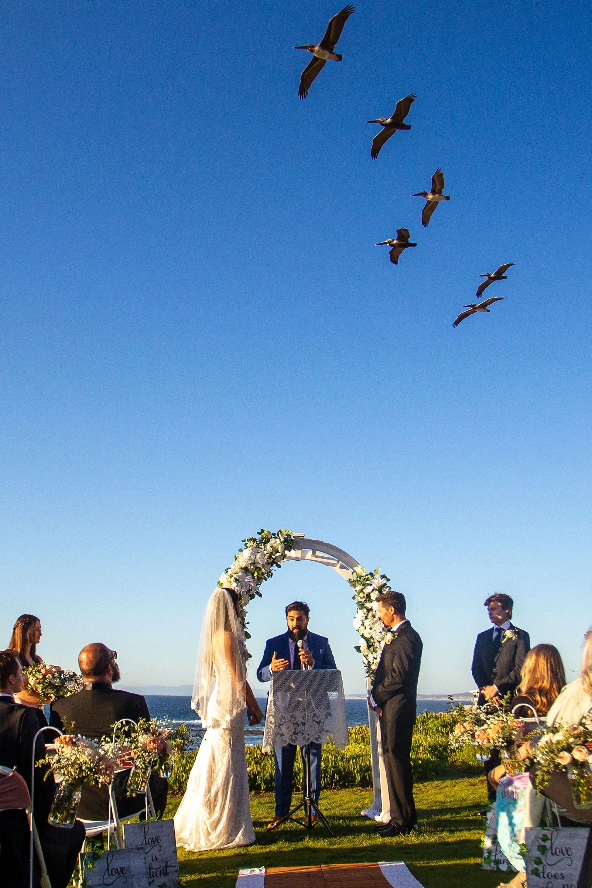 A wedding ceremony taking place outdoors, with a bride and groom standing under a floral arch next to an officiant, with guests seated on either side and overlooking the ocean, and birds flying in a clear blue sky.