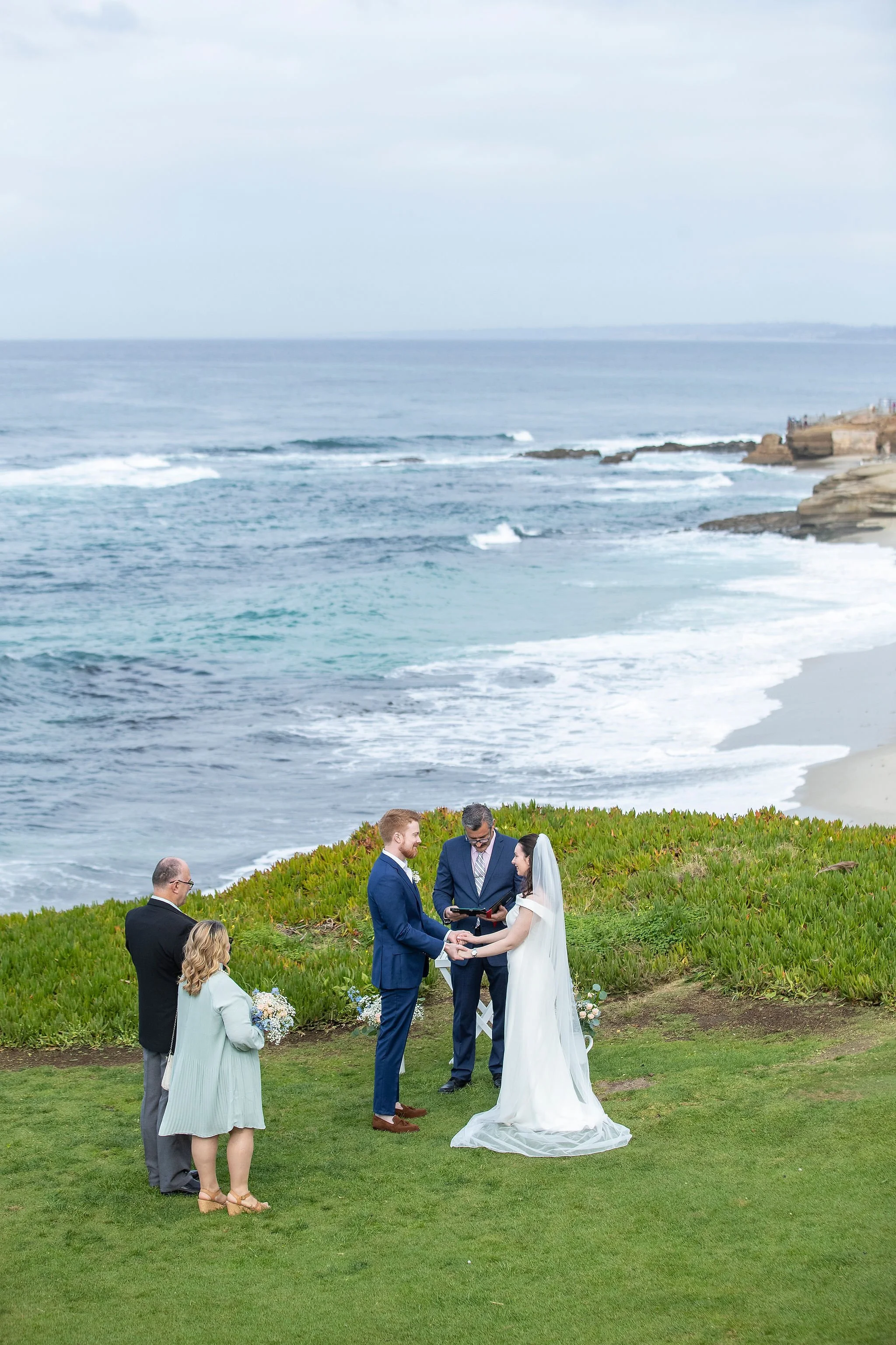 A wedding ceremony taking place on a grassy area near the ocean, with the bride and groom holding hands and exchanging vows, surrounded by officiant and guests, overlooking the beach and cliffs in the background.