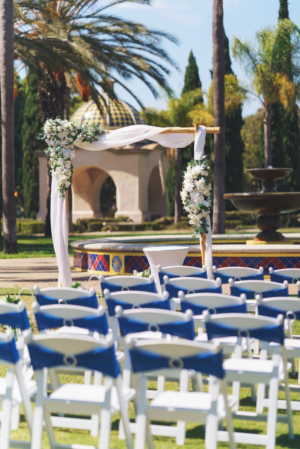Wedding ceremony setup outdoors with a floral arch, rows of white chairs with blue cushions, trees, and a fountain in the background.