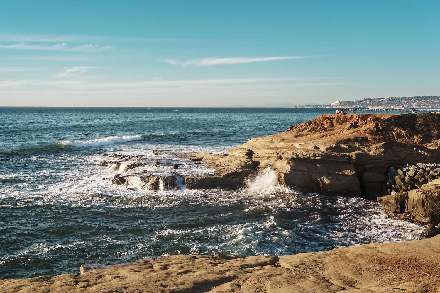 Rocky coastline with waves crashing and a pier in the distance against a clear blue sky.