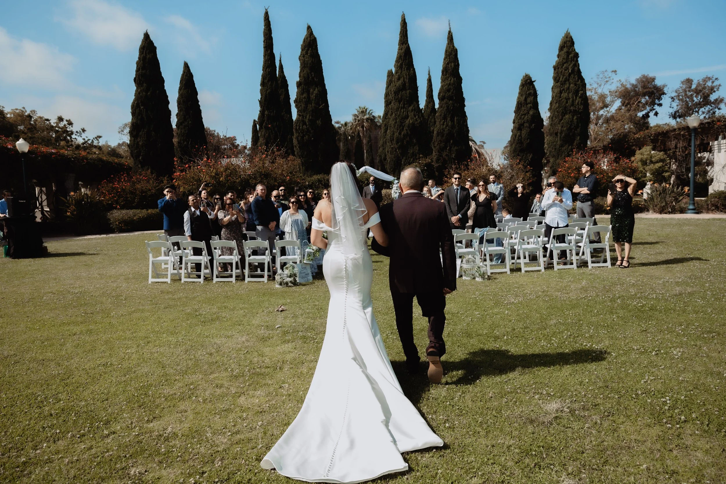 A bride and groom walking down an outdoor aisle during a wedding ceremony, with guests seated on either side and tall trees in the background.