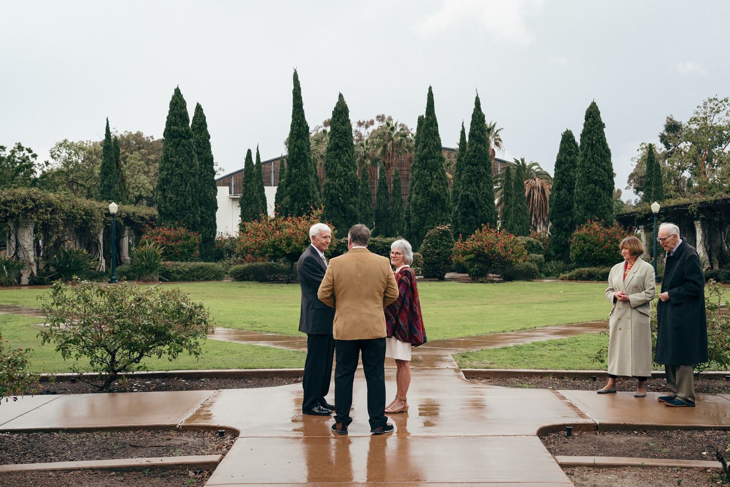 A group of five older adults standing outdoors on a rainy day, engaged in conversation on a wet paved walkway in a landscaped garden with tall cypress trees and flowering bushes in the background.