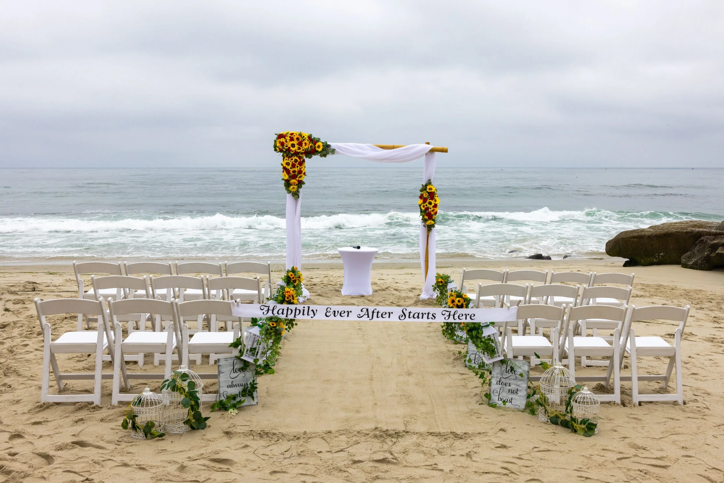 Beach wedding setup with white chairs, a decorated arch with sunflowers and white fabric, and a white sign that reads, 'Happily Ever After Starts Here.' The ocean and cloudy sky are in the background.