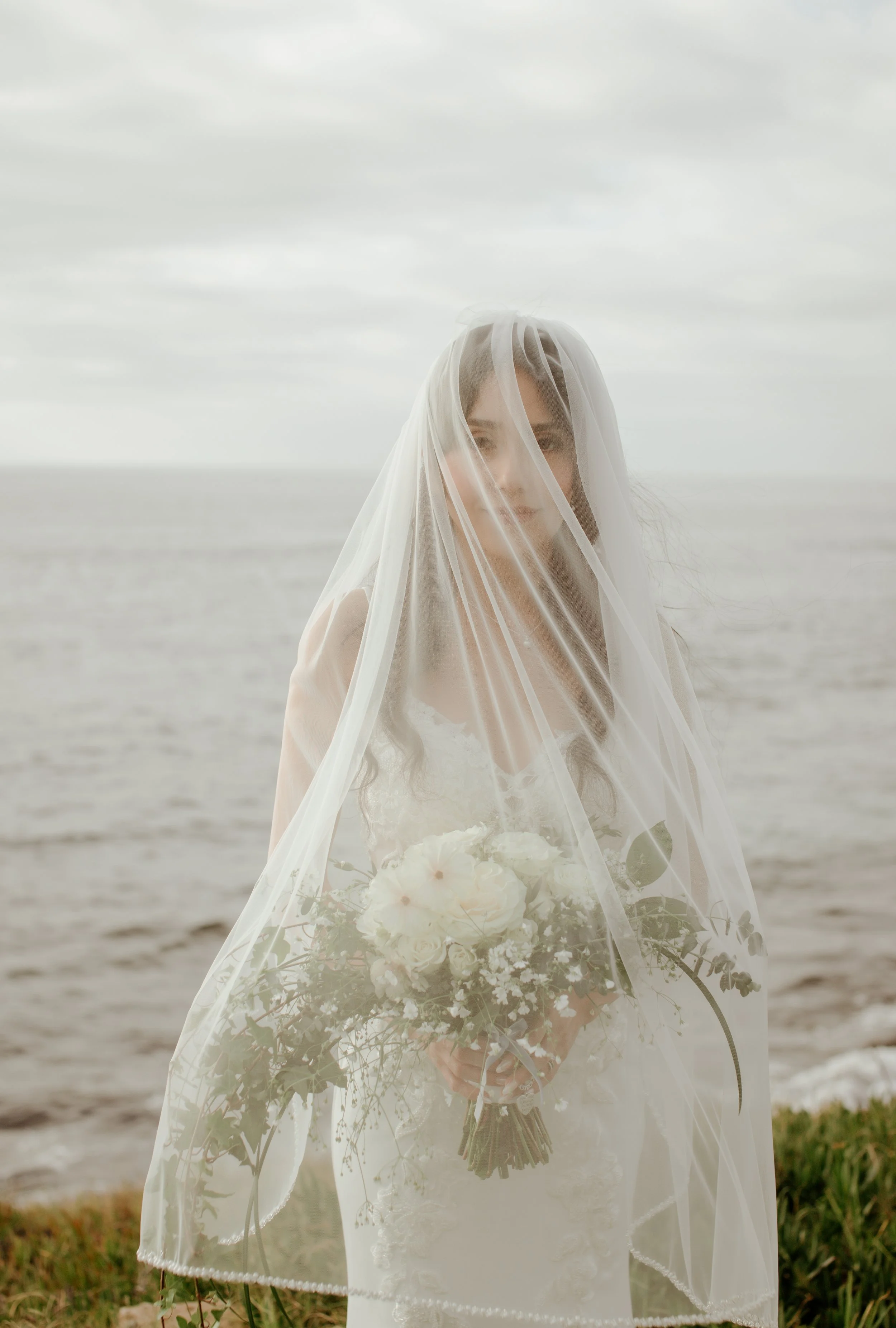 A woman dressed in a wedding gown, covered with a sheer veil, holding a bouquet of white flowers, standing on a beach with the ocean and cloudy sky in the background.