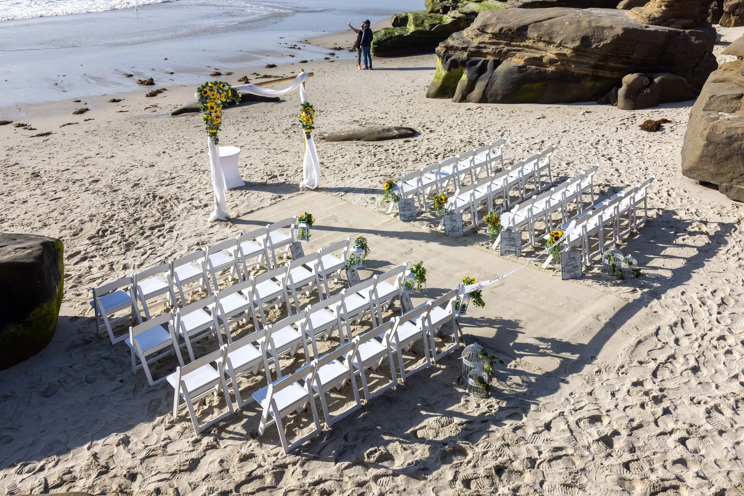 Beach wedding setup with white chairs, decorated with sunflowers, leading to an arch on the sand by the water. Two people are taking photos near large rocks; the ocean and sky are in the background.