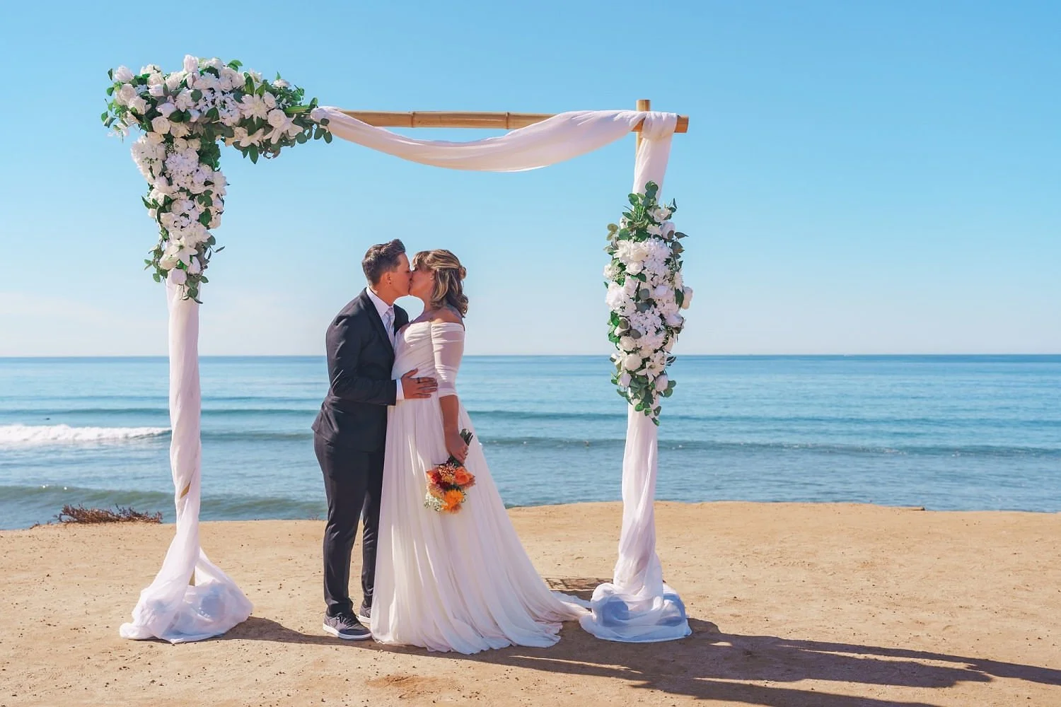 Couple in wedding attire sharing a kiss under a decorated arch on a beach with ocean and clear blue sky in the background.