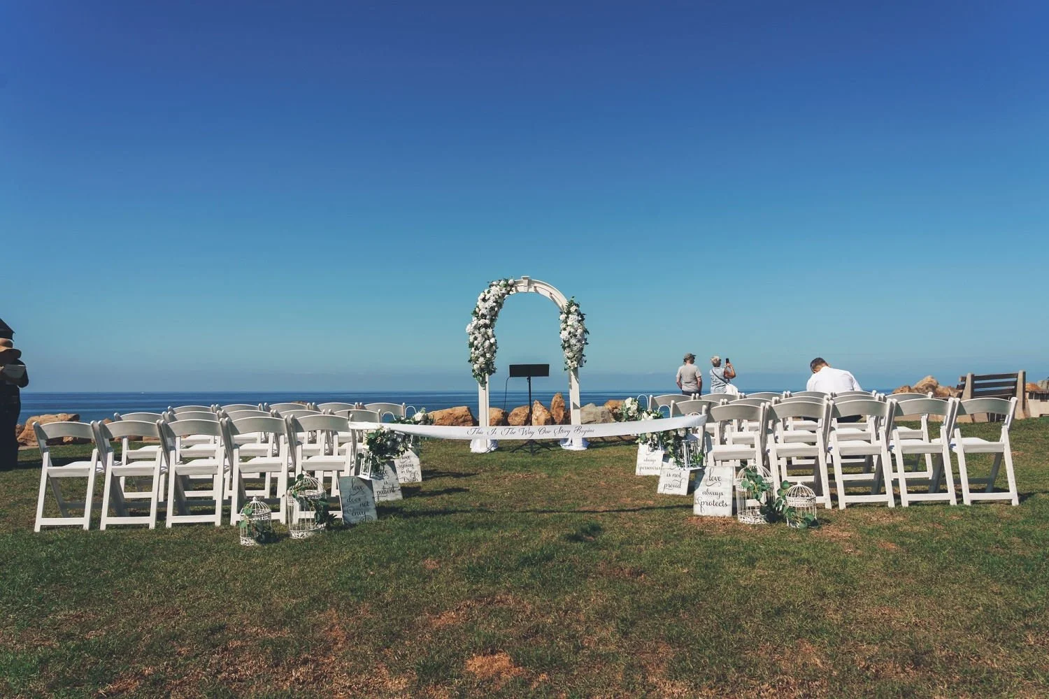 Wedding ceremony setup on a grassy area overlooking the ocean. White chairs are arranged facing an arch decorated with white flowers. Guests are seated or standing, with a few taking photos, under a clear blue sky.