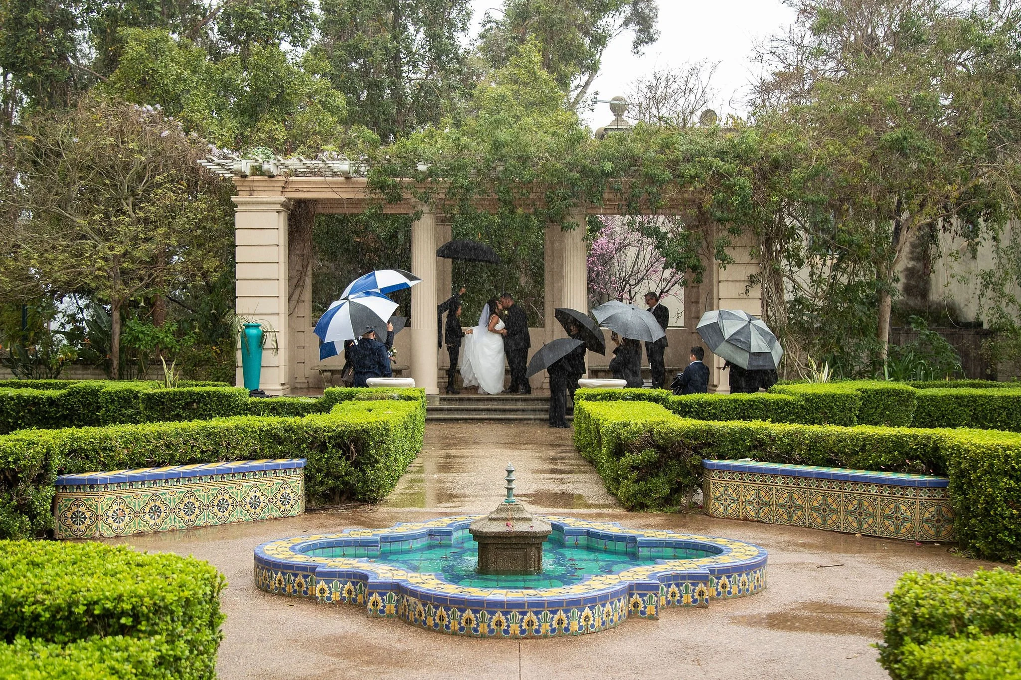 A wedding ceremony takes place in an outdoor garden under a classical archway with columns, surrounded by lush greenery and trees, during a rainstorm with umbrellas.