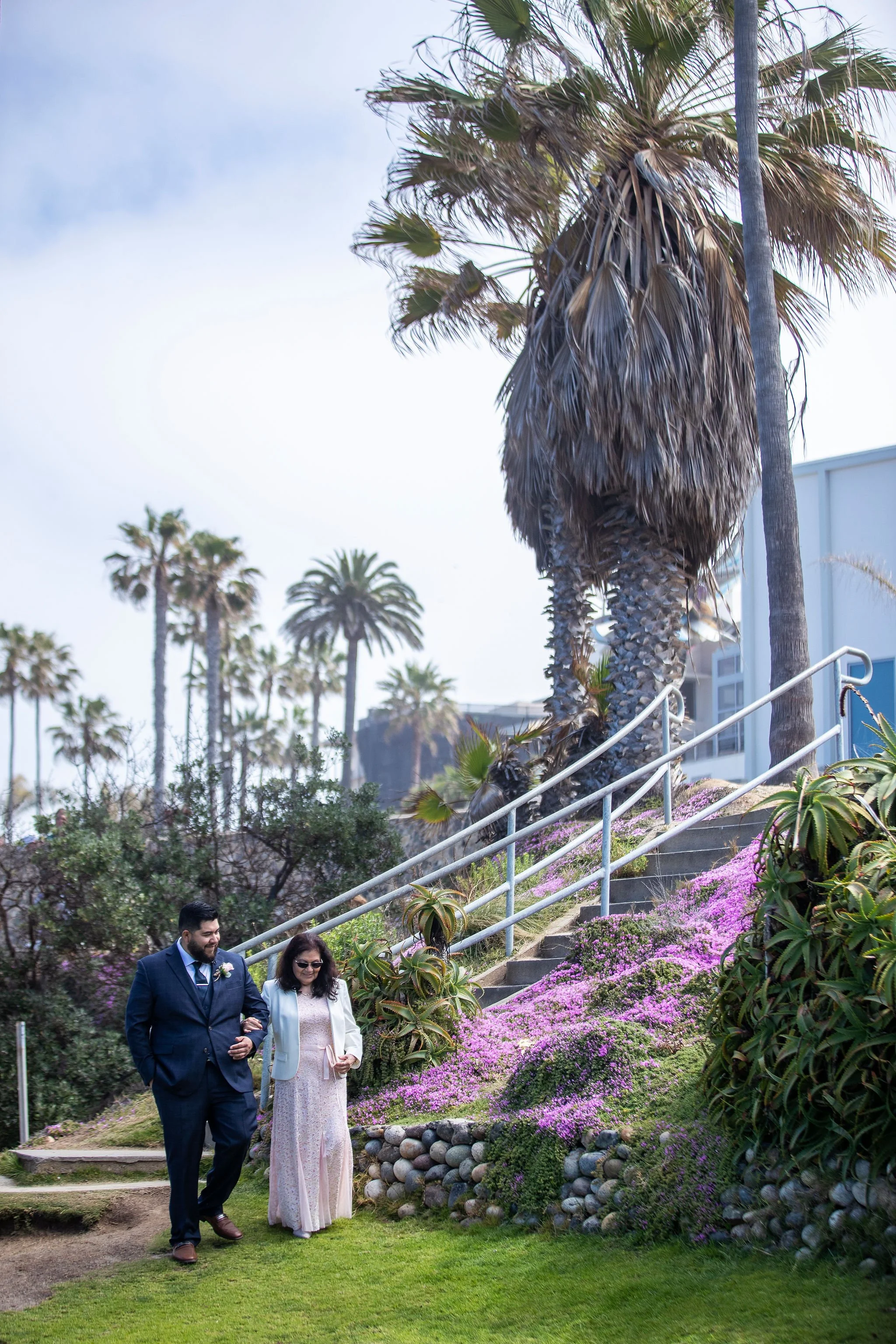 A couple walking down a garden path with purple flowers, alongside diverse shrubs and cacti, under tall palm trees, with a cloudy sky overhead.