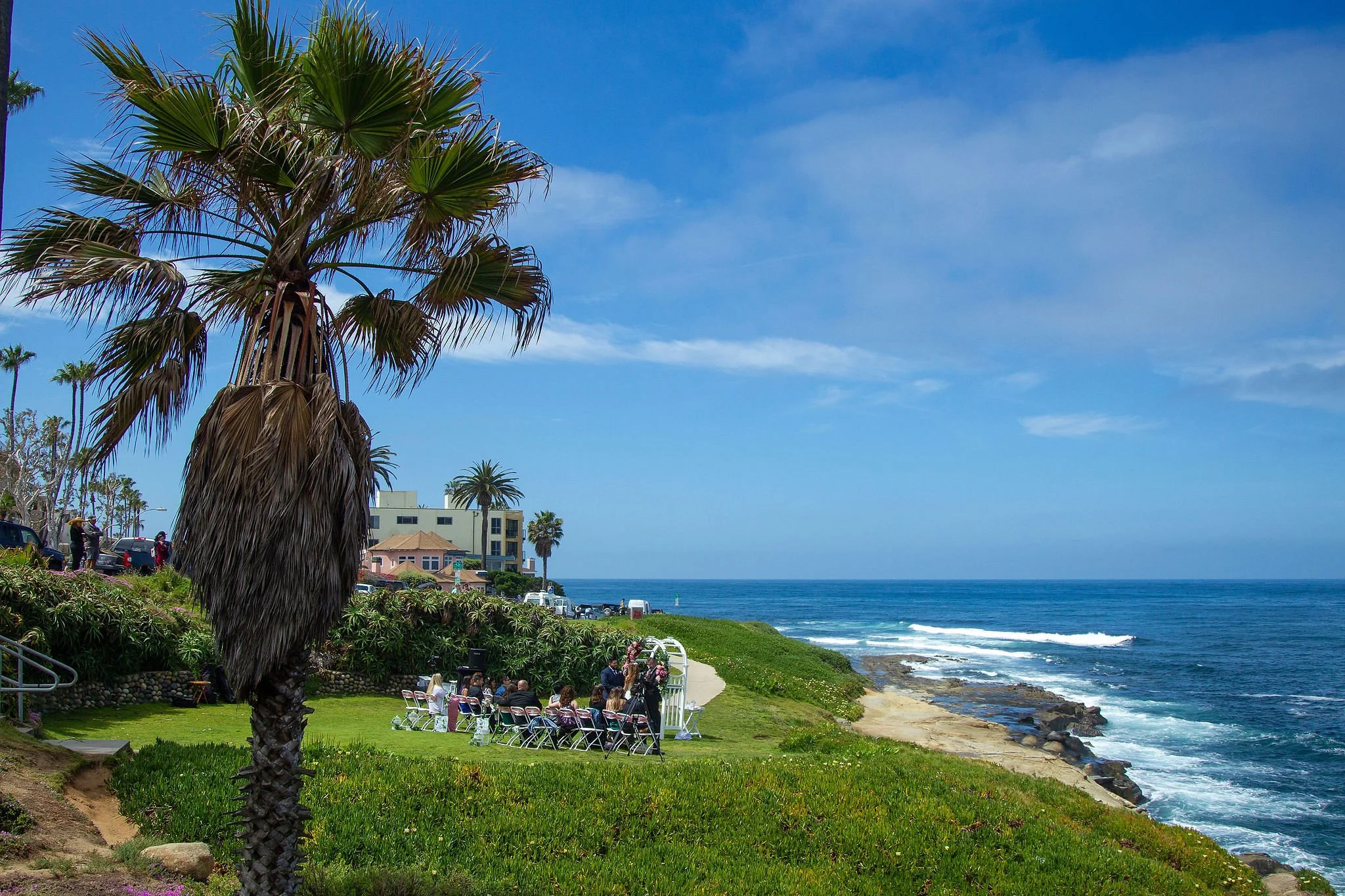 A group of people seated in chairs and standing under a white arch on a grassy area by the ocean, with sandy beach and rocks in the water. Palm trees and coastal buildings are in the background under a blue sky with some clouds.