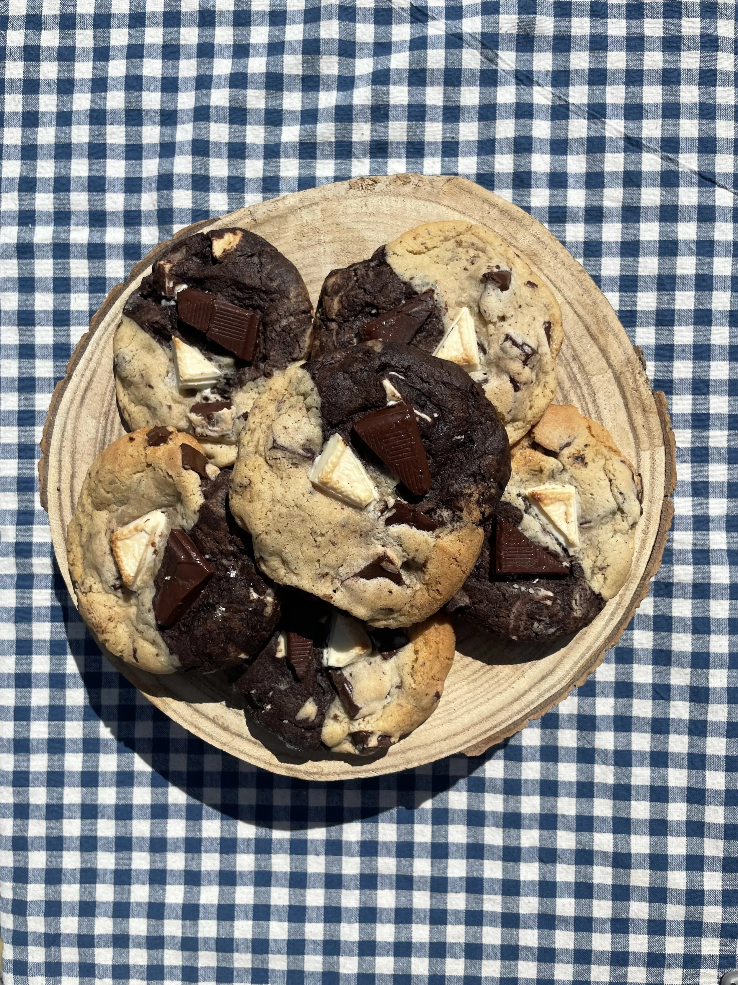 A wooden plate filled with homemade chocolate chip cookies topped with pieces of chocolate. The plate is on a blue and white checkered cloth.