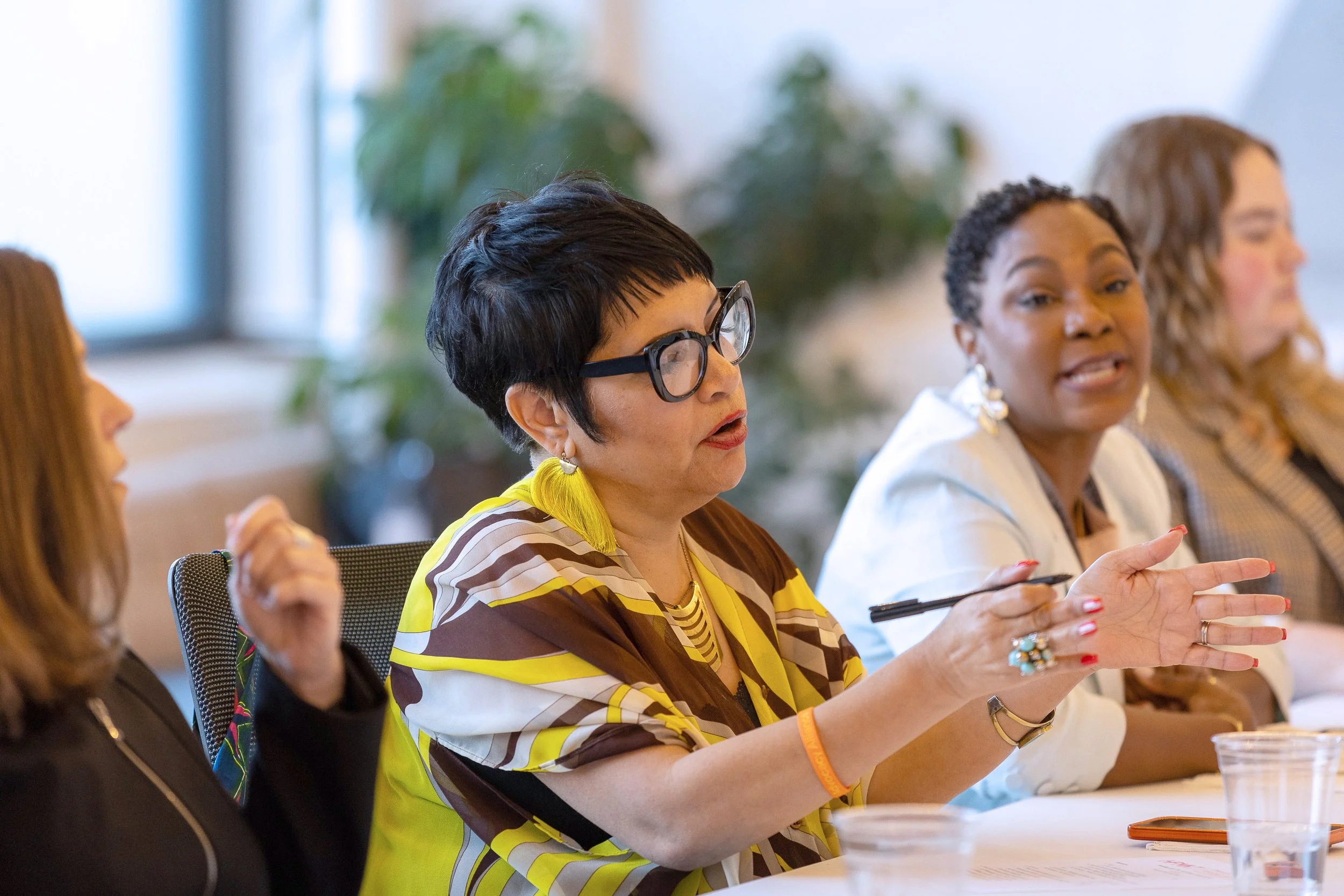 A diverse group of women sitting at a table in a meeting or discussion, with one woman in a striped yellow and brown shirt speaking and gesturing with her hands.