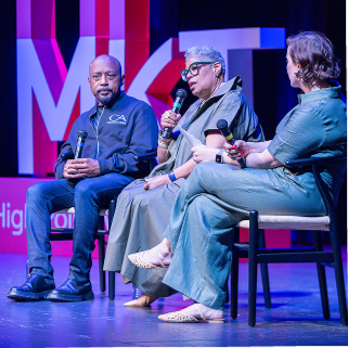 Three women sitting on stage at a panel discussion, with microphones in their hands, in front of a backdrop with the letters 'M'.