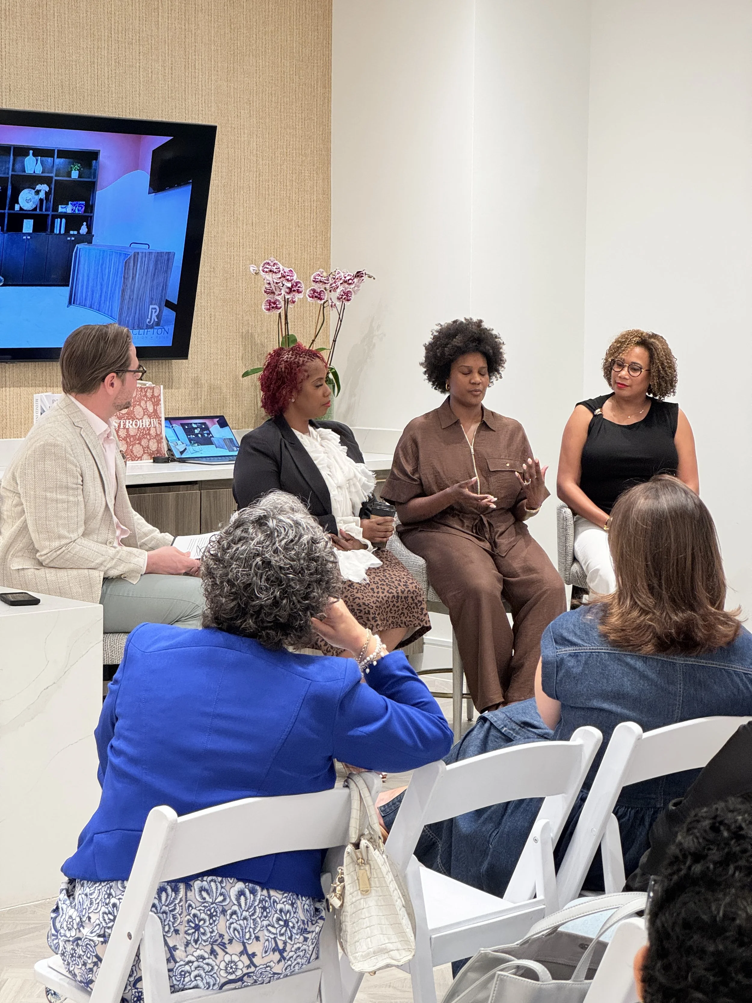 A panel discussion with four women seated in front of an audience. One woman is speaking with her hands, and the others are listening. The background includes a television screen, a shelf with decorative items, and an orchid plant. Audience members are sitting on white chairs.