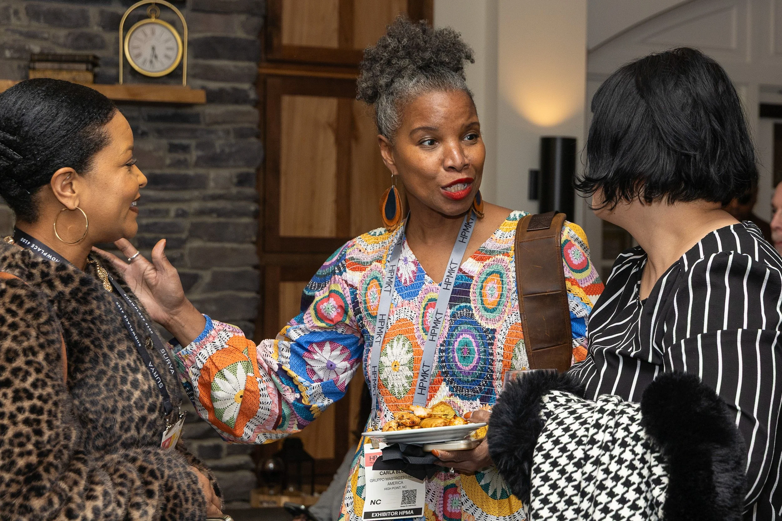 Three women engaged in conversation at a social gathering, with one woman in colorful attire holding a plate of food. The background features a stone wall, a clock, and wooden shelves.