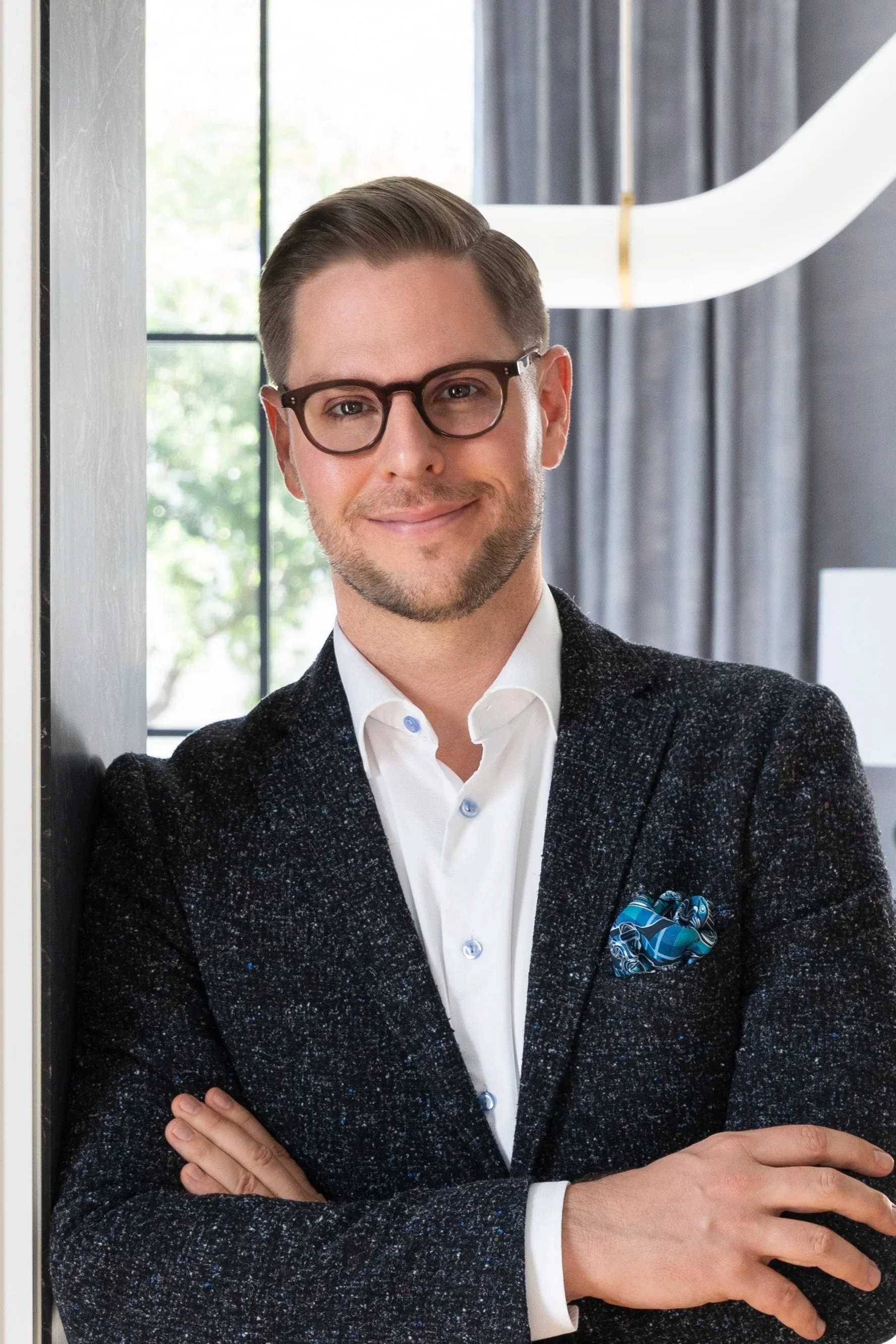 A man with glasses wearing a white shirt and a dark patterned blazer, standing with arms crossed in an indoor setting with a large window and curtains in the background.
