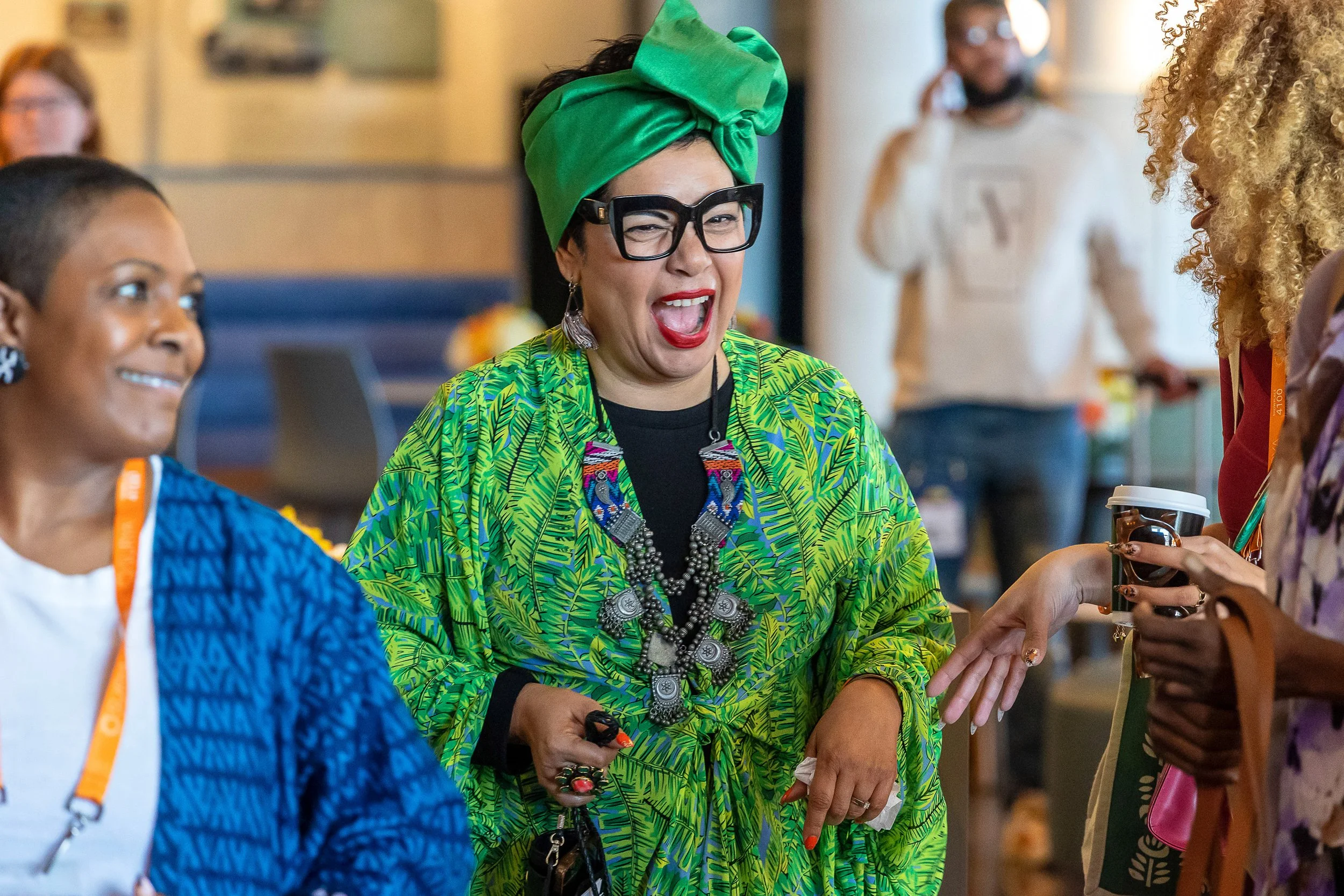 A woman wearing a bright green headscarf, large black glasses, and colorful tropical-patterned clothing, smiling and engaging with others at a social event.