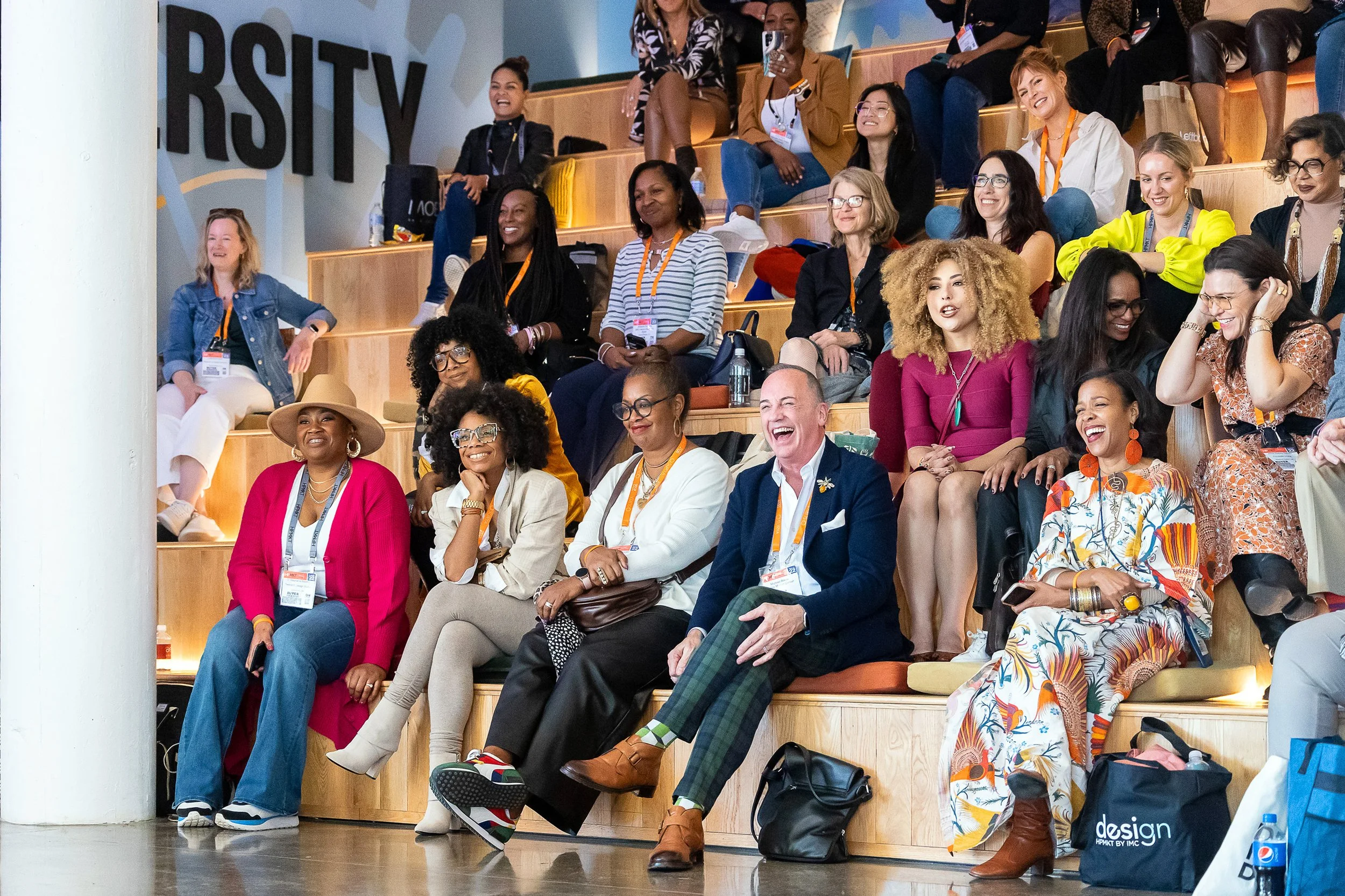 A group of diverse women and men sitting on tiered wooden seats at a conference, laughing and smiling, with some taking photos and others engaging in conversation, inside a modern venue with a large