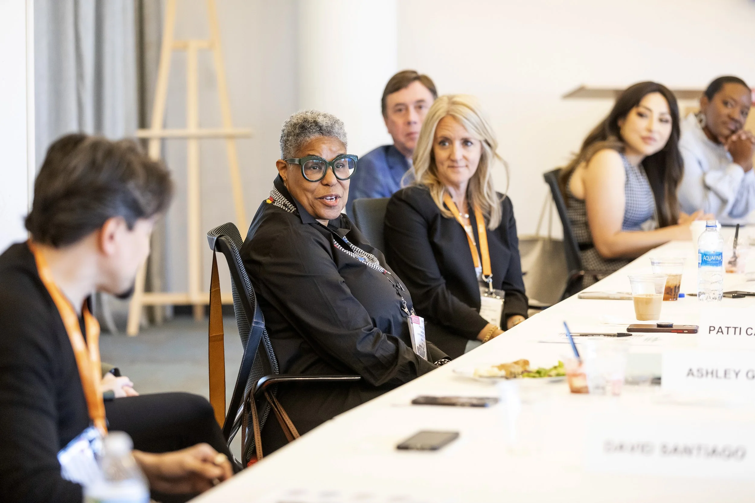 A diverse group of five women and one man sitting at a conference table during a meeting. The woman in the forefront has gray hair and glasses, and the others are listening attentively. There are drinks and snacks on the table.