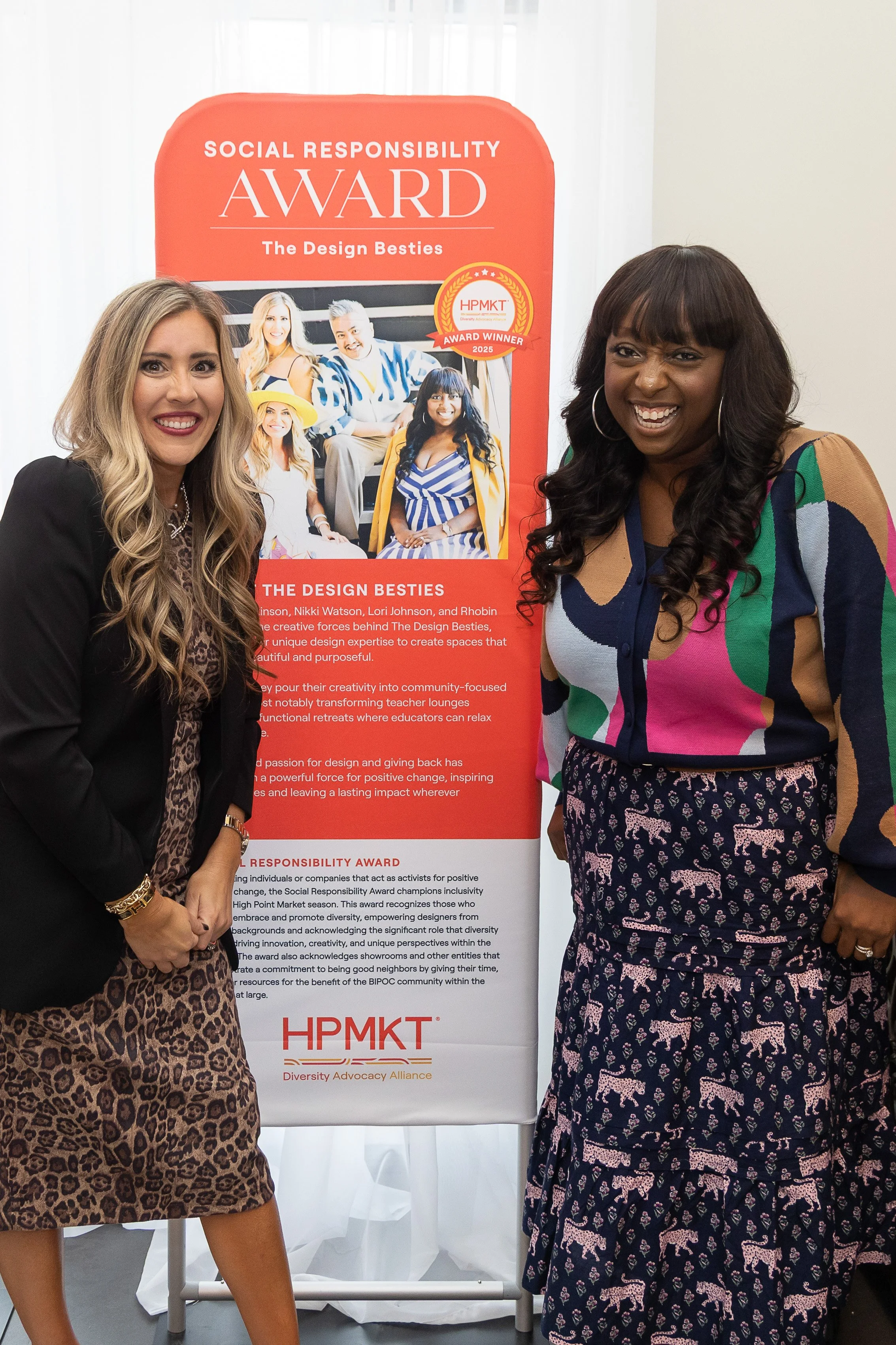Two women standing in front of a red poster that reads 'Social Responsibility Award'. They are smiling and appear to be at an event. The woman on the left has long blonde hair and is wearing a black blazer over a leopard print dress. The woman on the right has long dark hair, large hoop earrings, and is wearing a colorful top and a dark skirt with pink panther print.