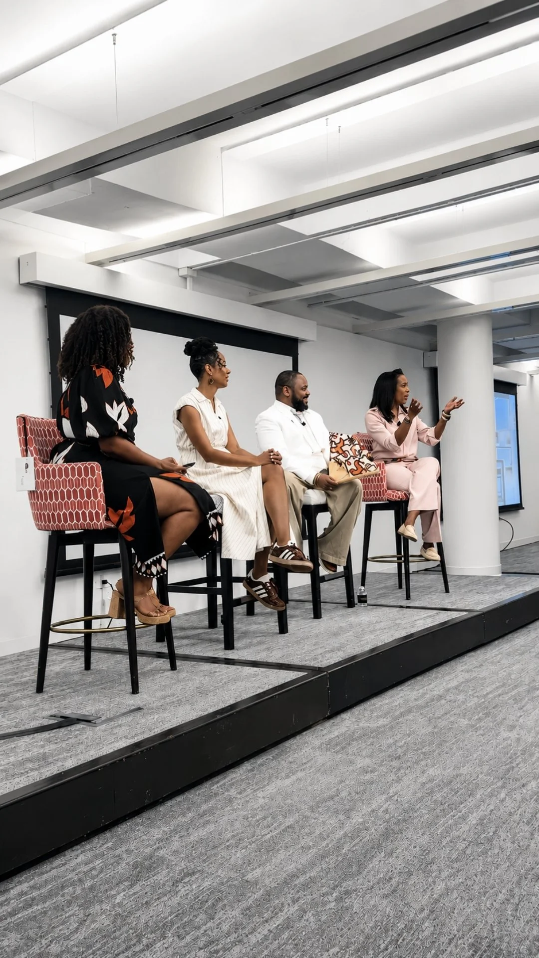 Four people sitting on high chairs on a stage, engaged in a panel discussion or presentation in a modern conference room.