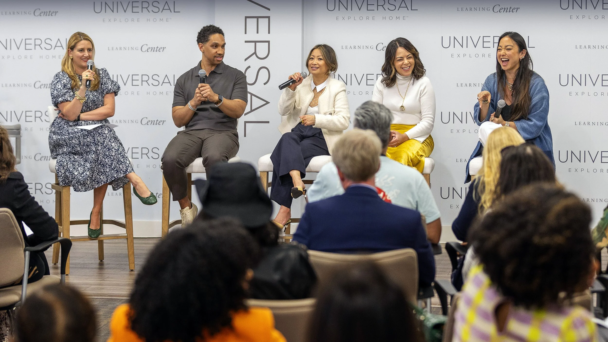 A diverse group of five women and one man having a lively discussion on stage at a panel discussion during an event at Universal Explore Home Learning Center, with an audience in front.