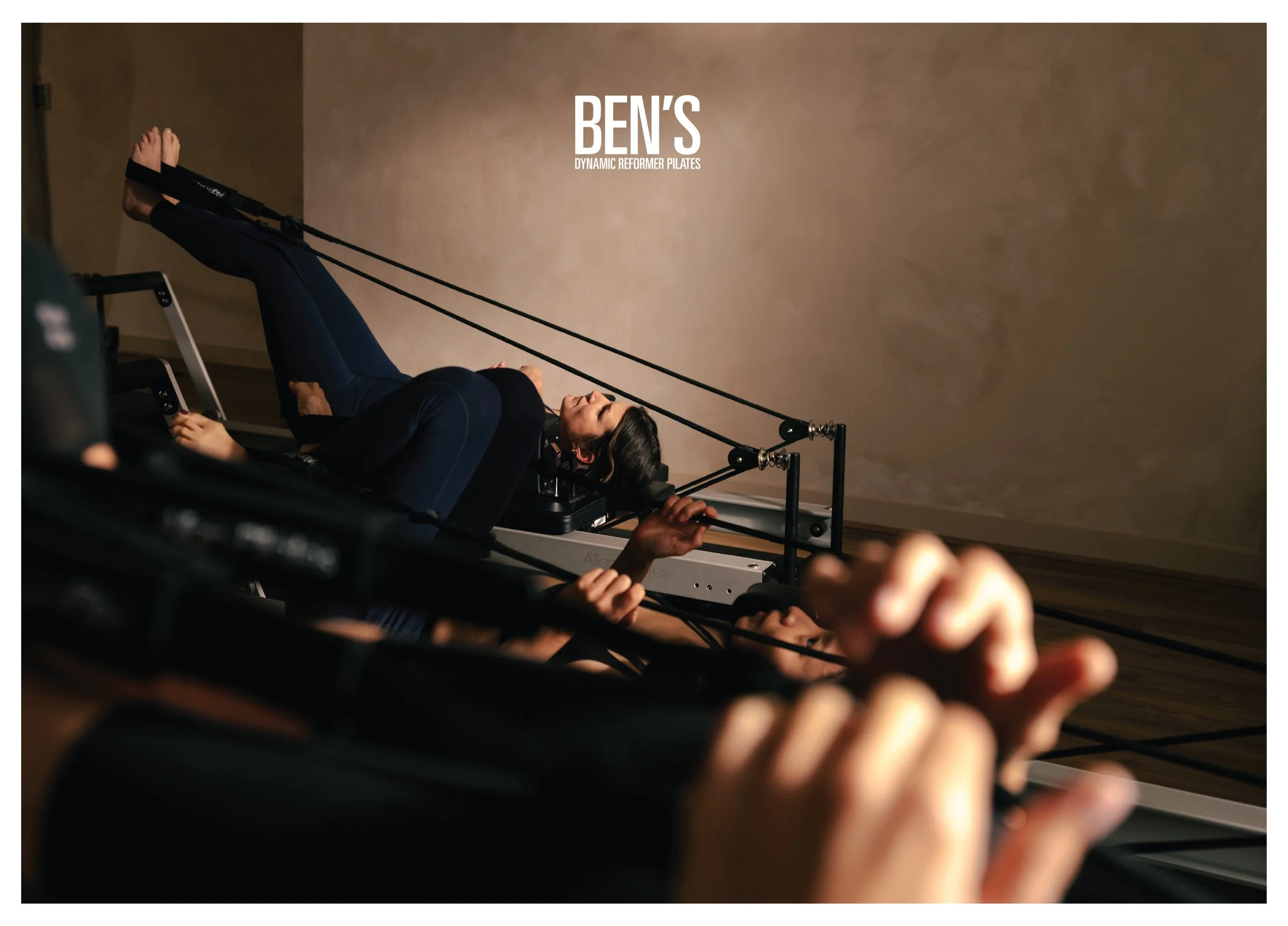 A woman lying on a Pilates reformer machine with resistance straps, focusing on her exercise, in a studio with neutral-colored walls.