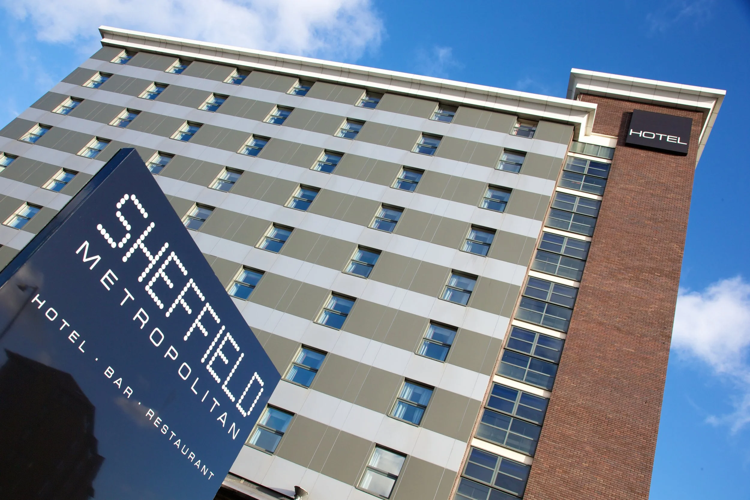 Modern multi-story hotel building with a mix of beige, gray, and brick exterior, and a large black sign with white text reading "Hotel" on the upper right. A blue directional sign in the foreground points to different services like restaurant, bar, a