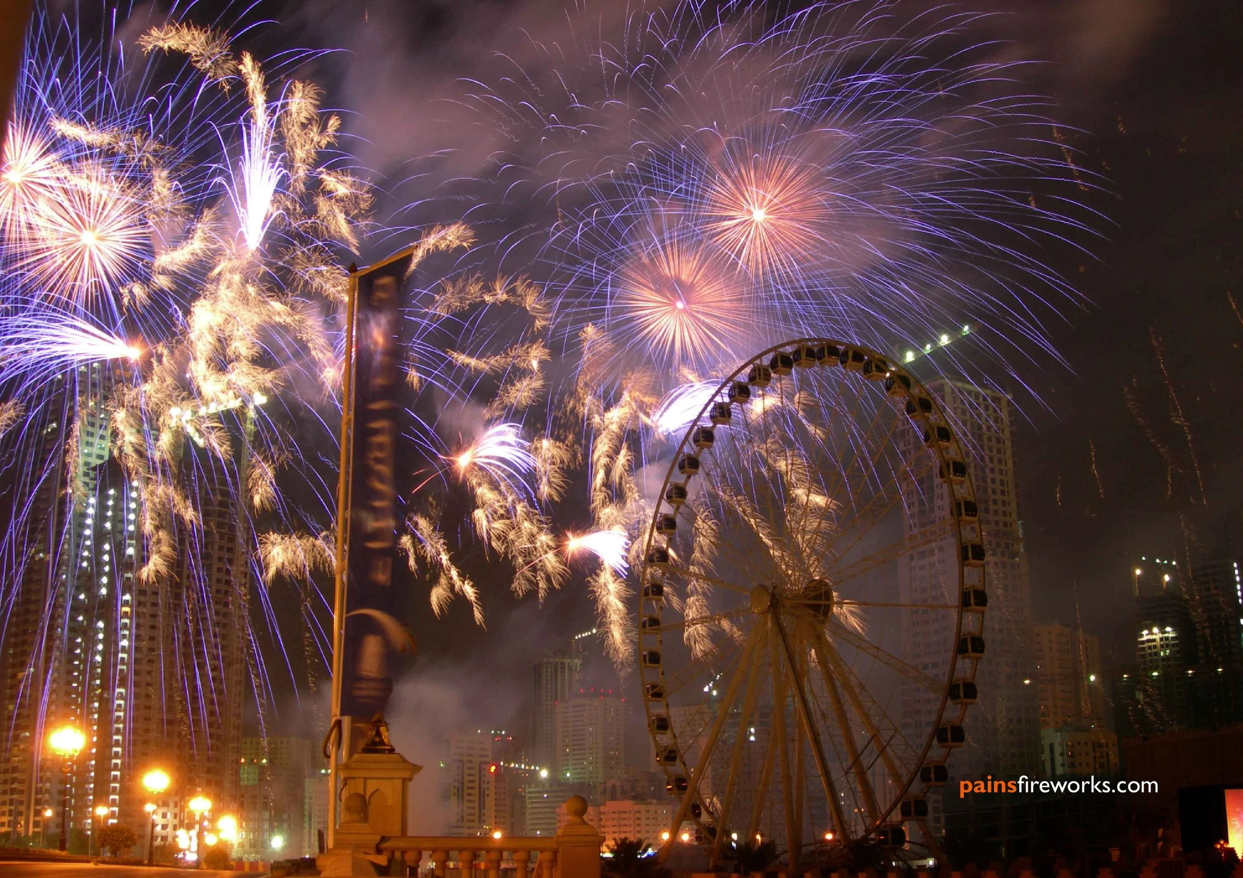 Fireworks lighting up the night sky above an amusement park with a Ferris wheel, tall buildings, and streetlights in the background.