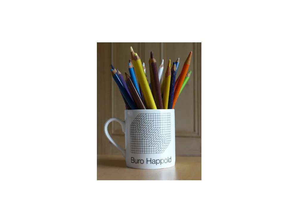 A white mug filled with colorful pencils, placed on a wooden surface with a wooden background. The mug has a geometric design and the text 'Buro Happold' printed on it.