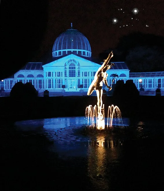 Nighttime view of a glass conservatory illuminated in blue light, with a lit fountain sculpture of a ballerina in the foreground and starry sky above.