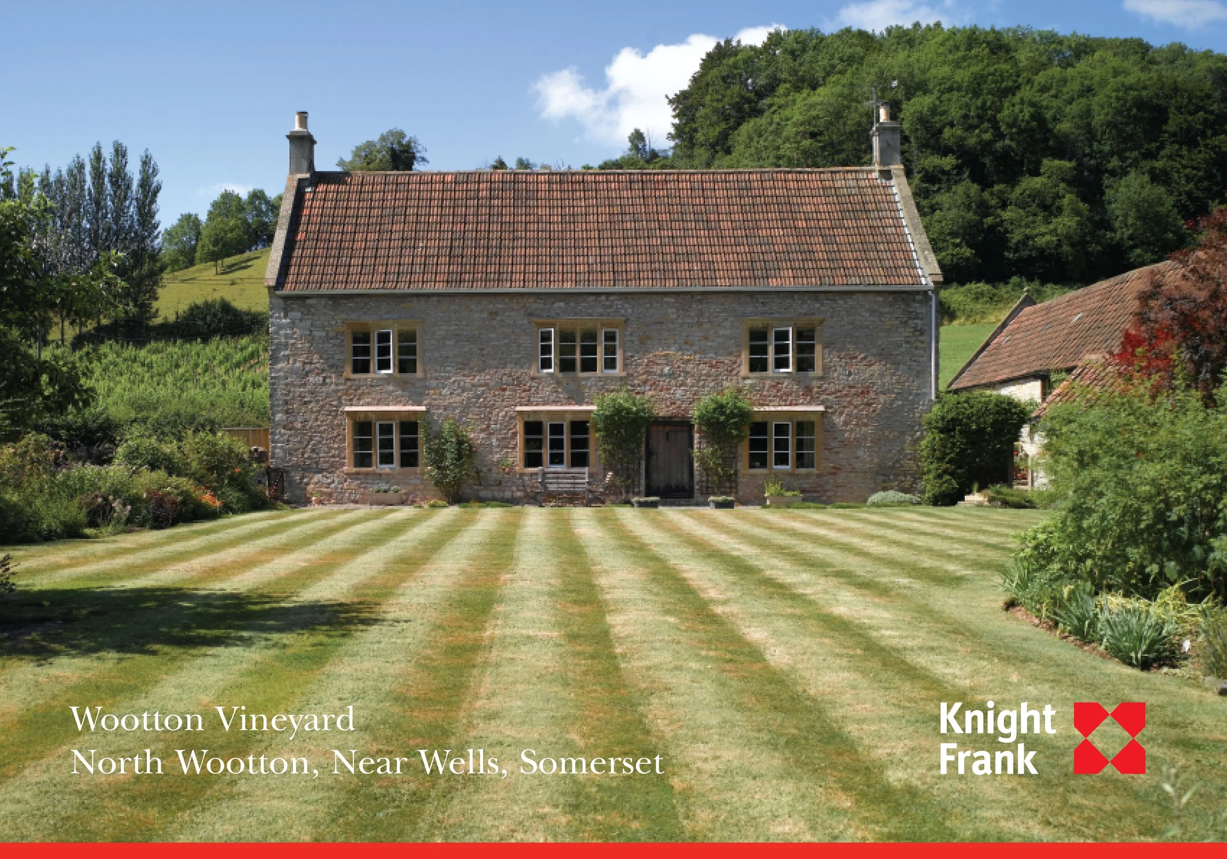 A stone house with multiple windows and a door, surrounded by a well-maintained lawn and garden, with trees and hills in the background under a partly cloudy sky.