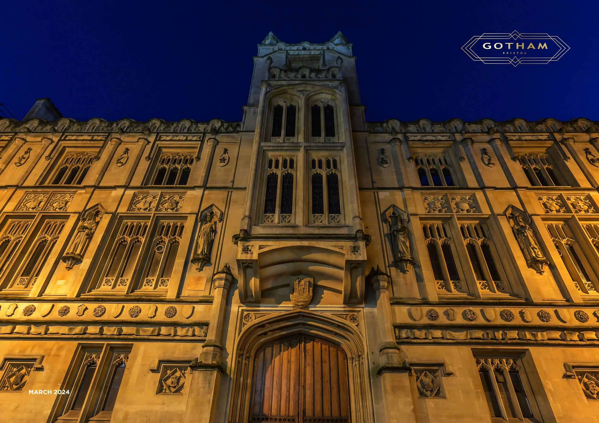 Nighttime view of a historic gothic-style building with illuminated architectural details, including arched windows, sculptures, and a central tower with a clock or decorative element, under a dark sky. The image is tagged with 'Gotham Bristol' in th
