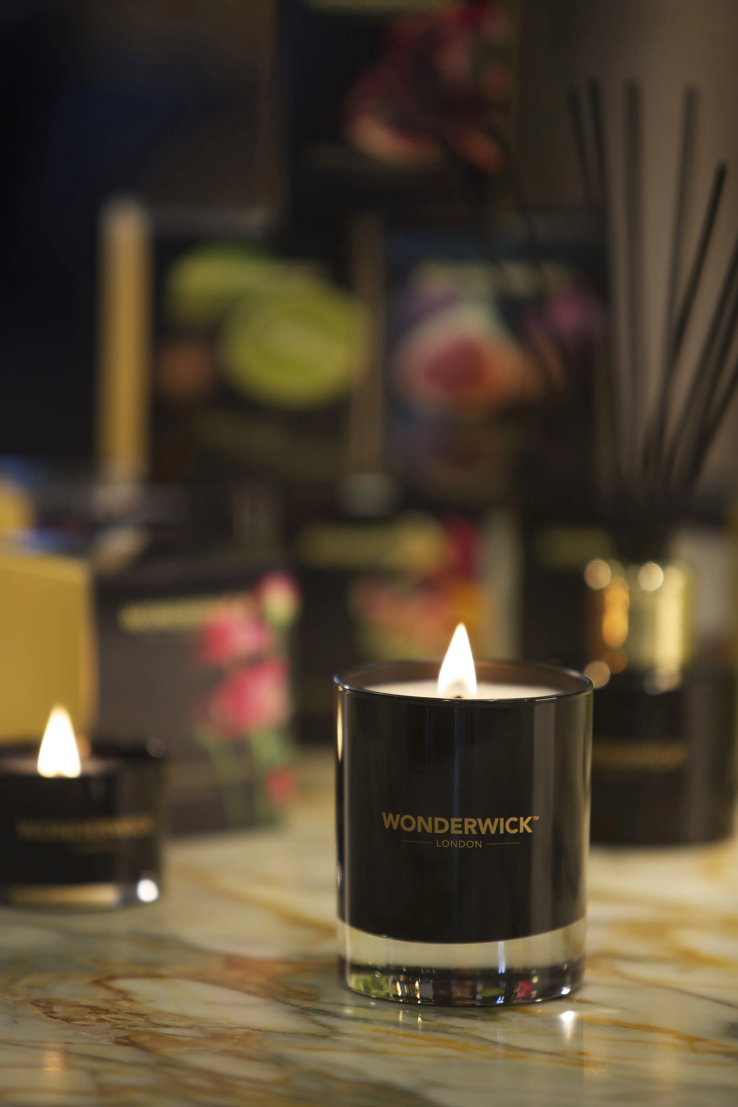 A burning candle in a black container labeled WONDERWICK London on a marble surface, with blurred candles and a colorful floral arrangement in the background.
