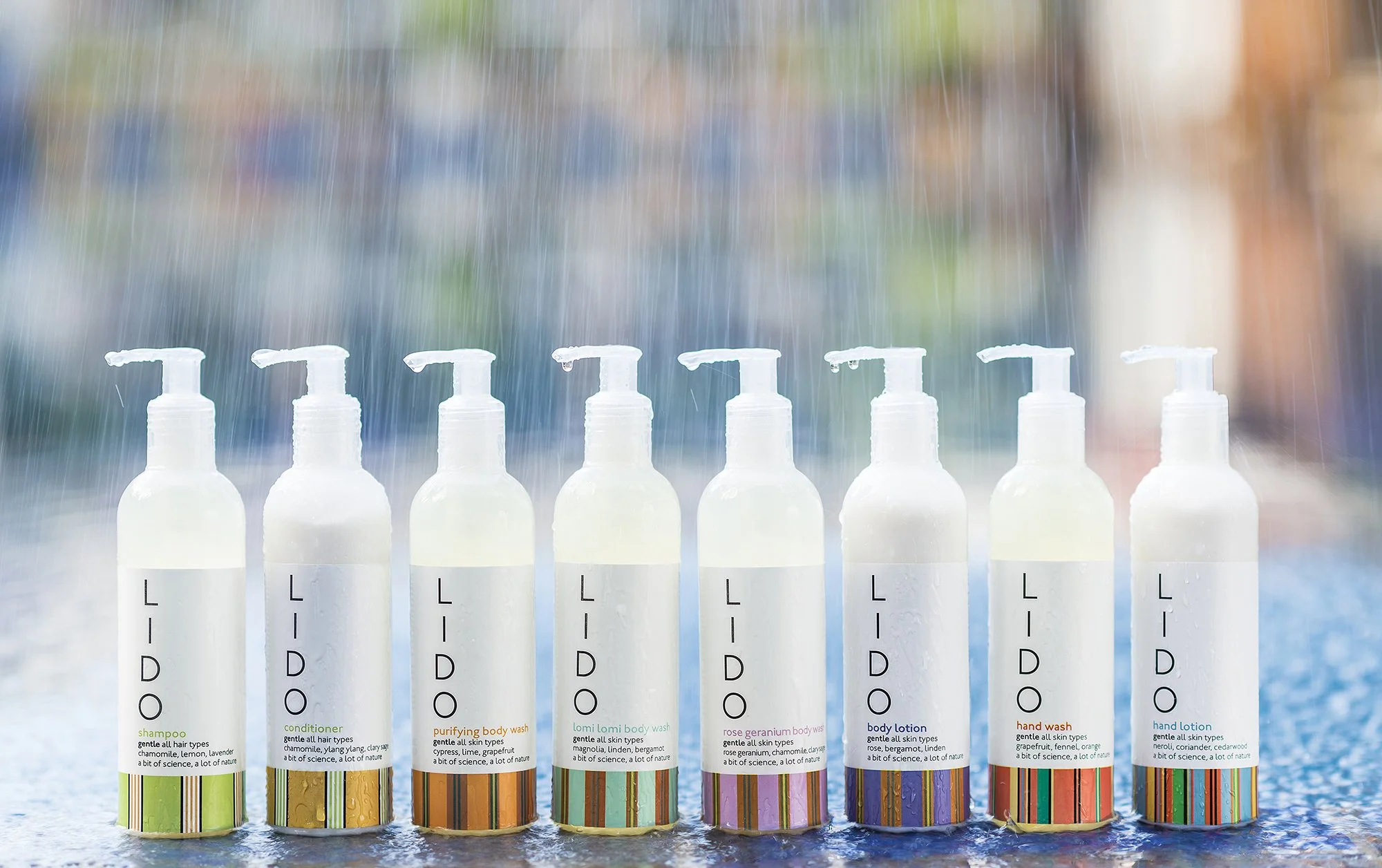 A row of eight white bottles with pump dispensers labeled for personal care products, placed on a wet surface with water splashes, with a blurred colorful background.
