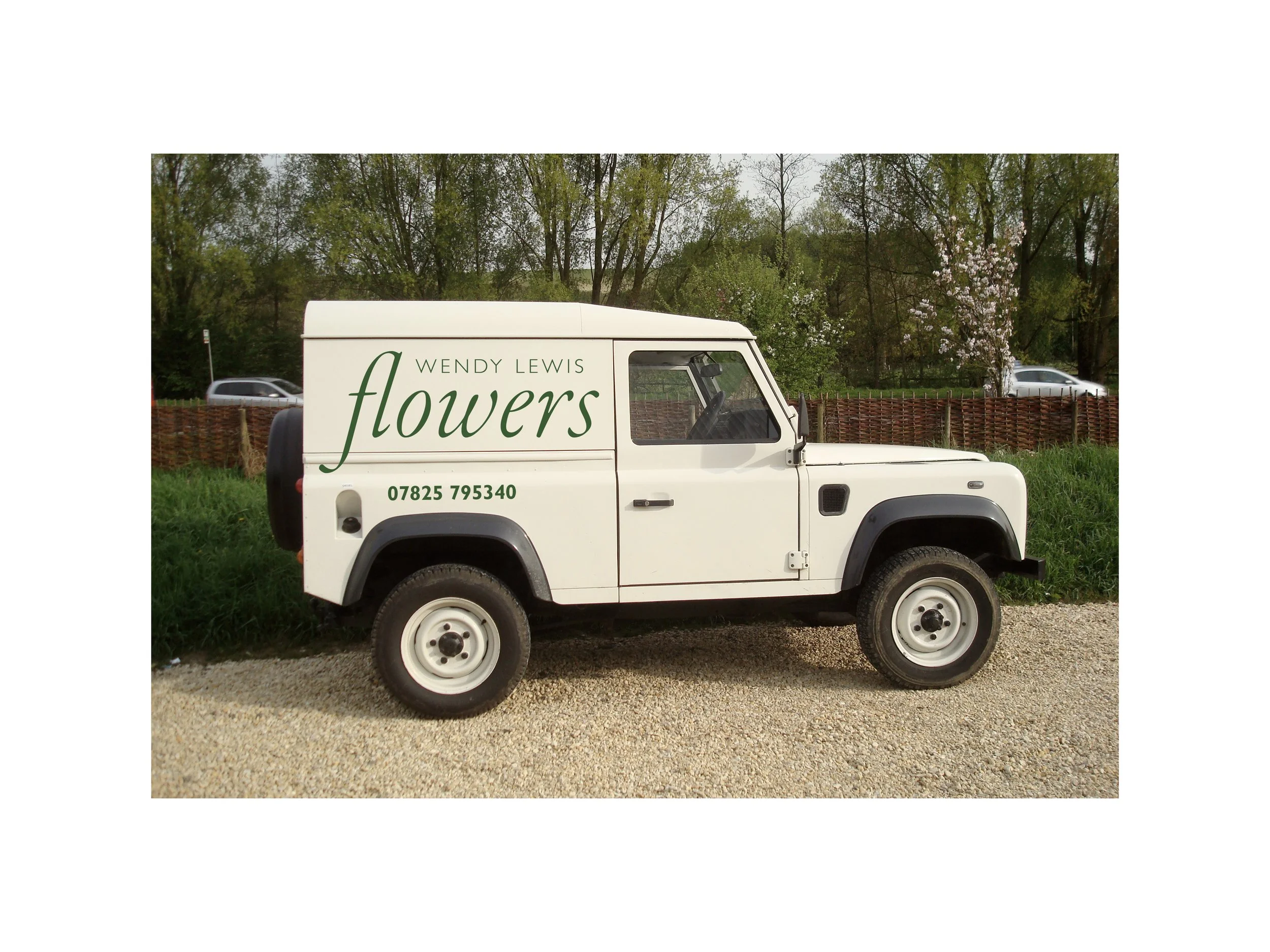 A white delivery van with green lettering for Wendy Lewis Flowers, parked on a gravel surface with trees and cars in the background.