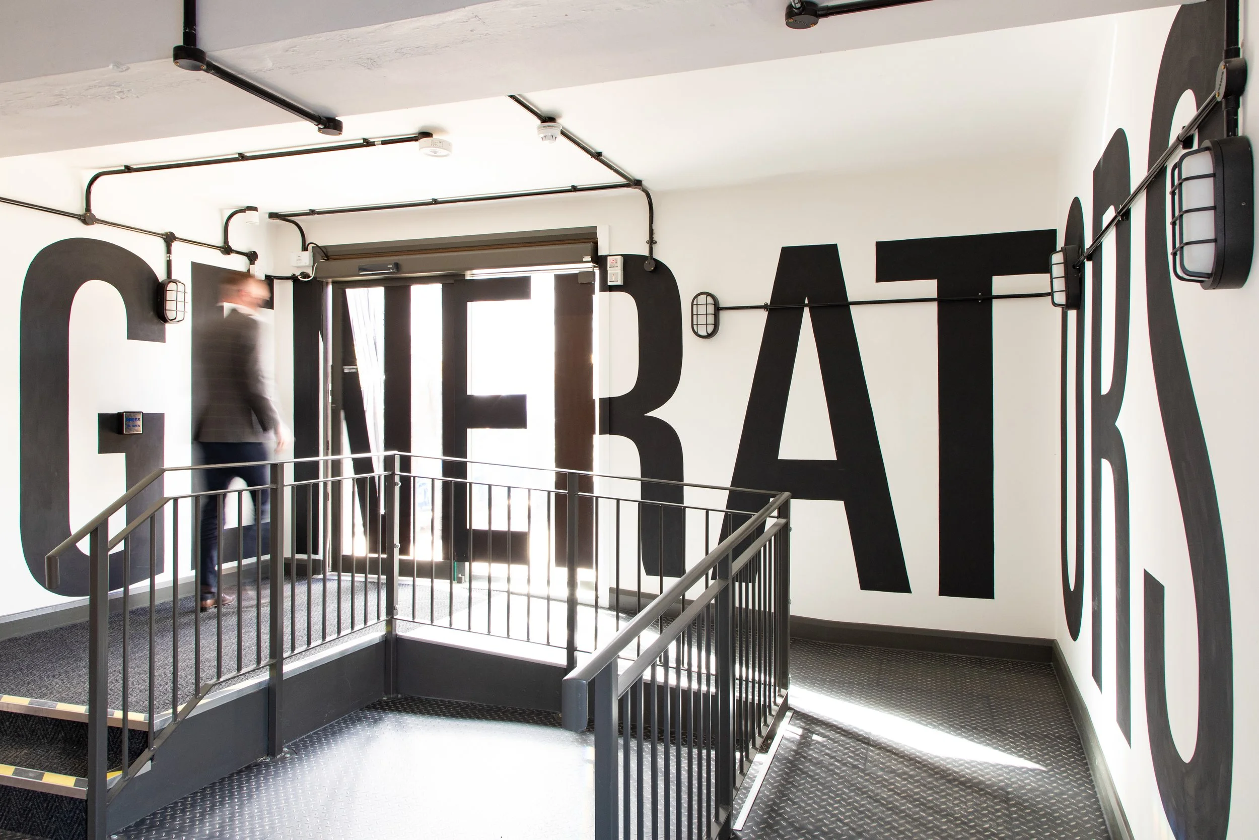Interior of a modern building with a staircase, black lettering on white walls, industrial-style piping on the ceiling, and a blurred person walking.