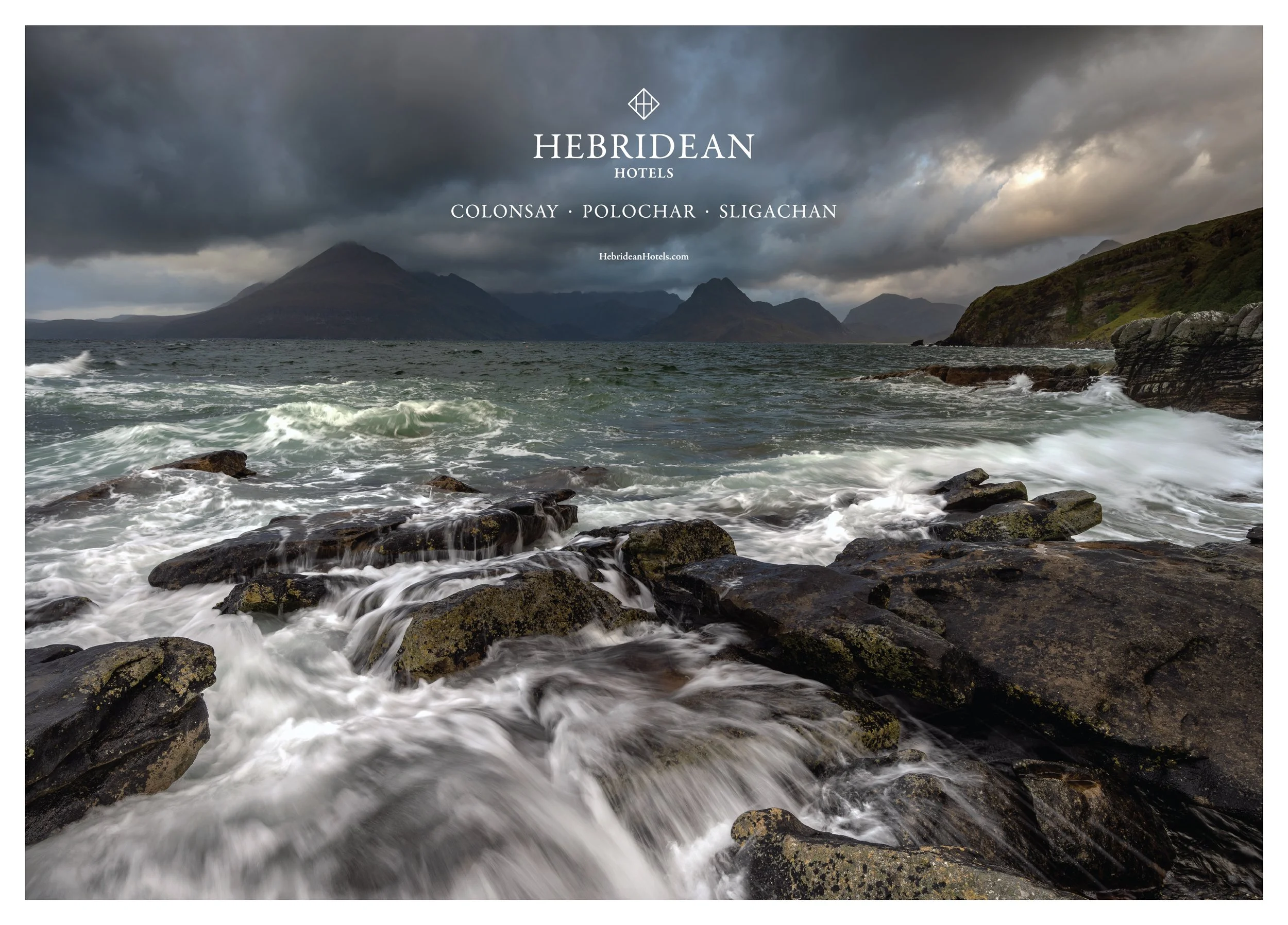 A rocky coastline with water splashing over rocks and mountains in the distance under a cloudy, stormy sky.
