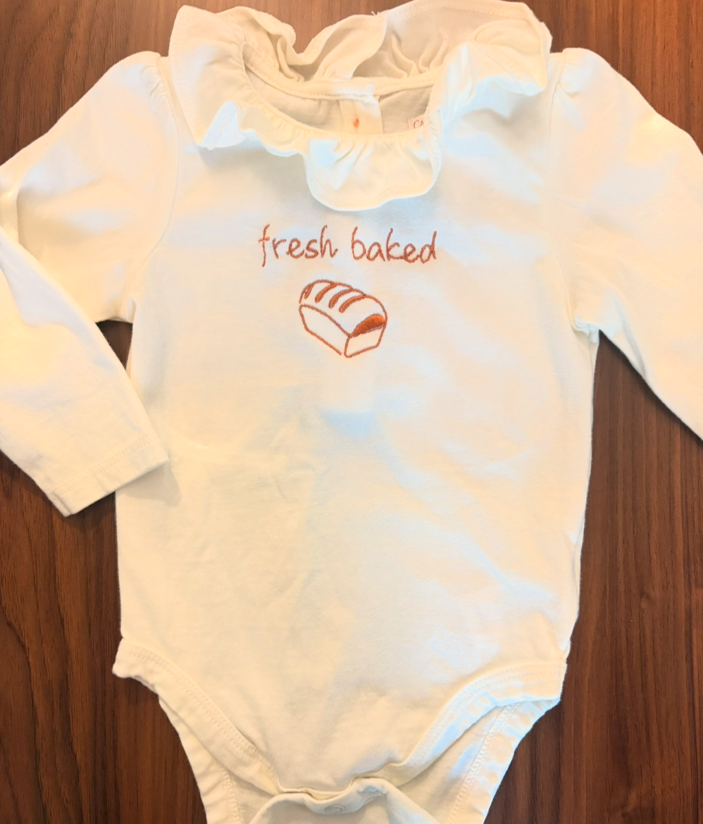 Cream-colored long sleeve baby onesie with ruffled collar, embroidered text 'fresh baked', and a baking pan with bread illustration.
