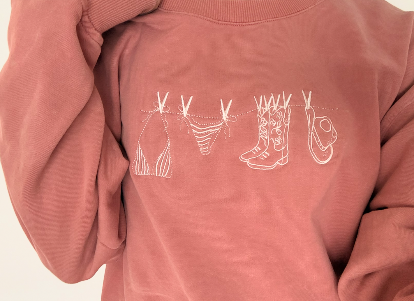A person wearing a pink hoodie with a white embroidered design of clothing and shoes hanging on a clothesline.