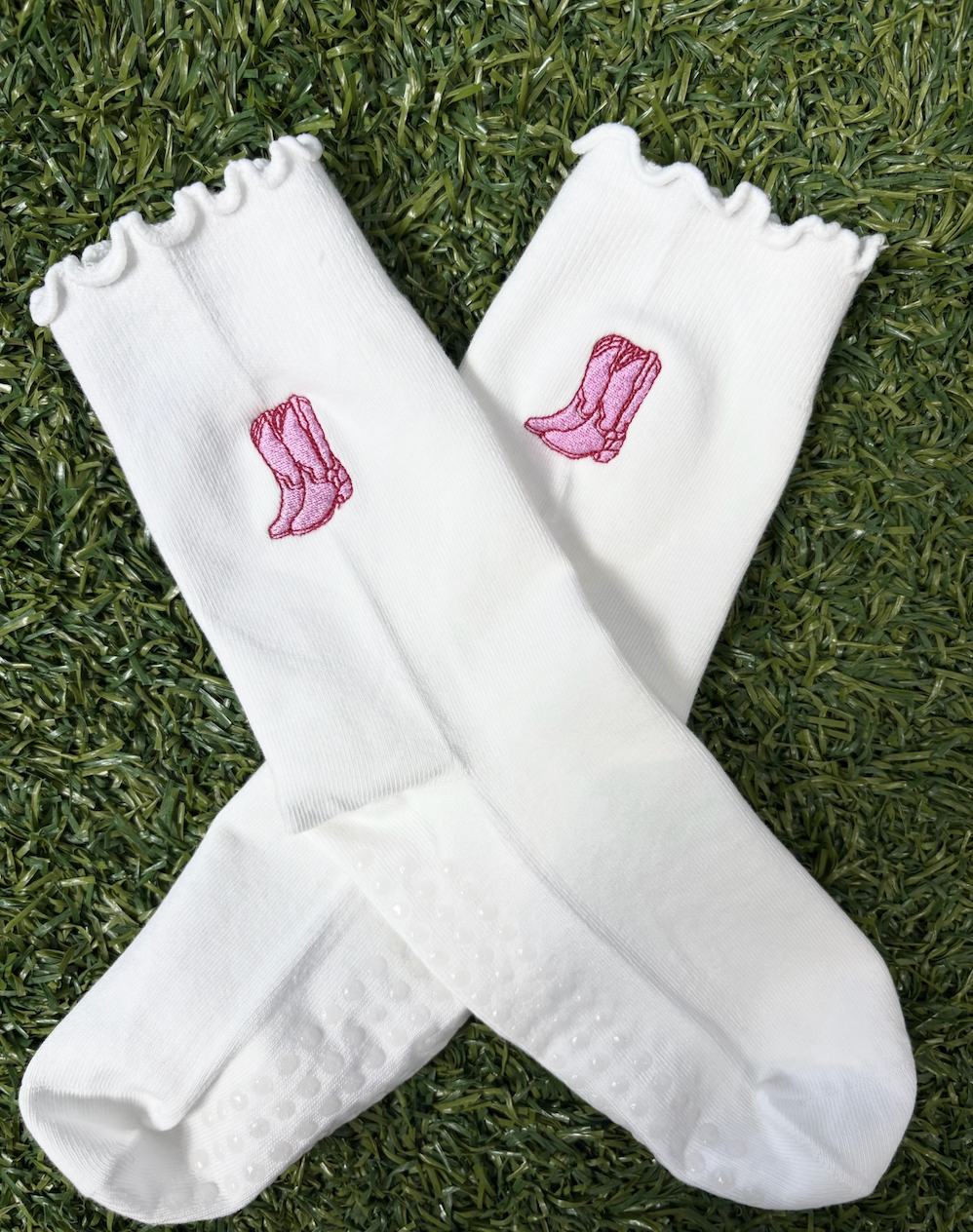 Pair of white socks with pink cowboy boots embroidered on each, laid on green grass.