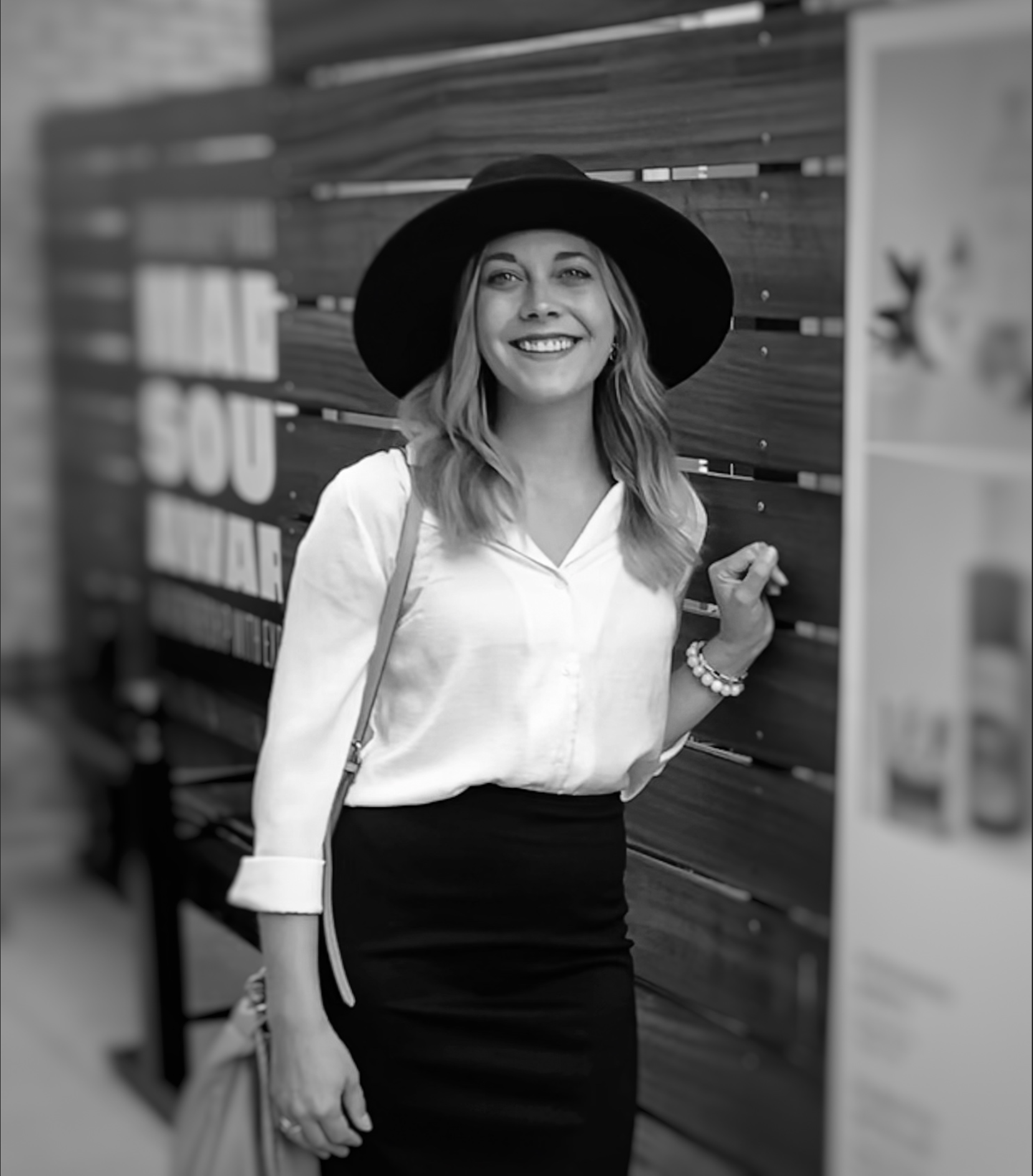 A smiling woman wearing a wide-brimmed hat and a white blouse, standing in front of a wooden fence with blurred text and design elements.