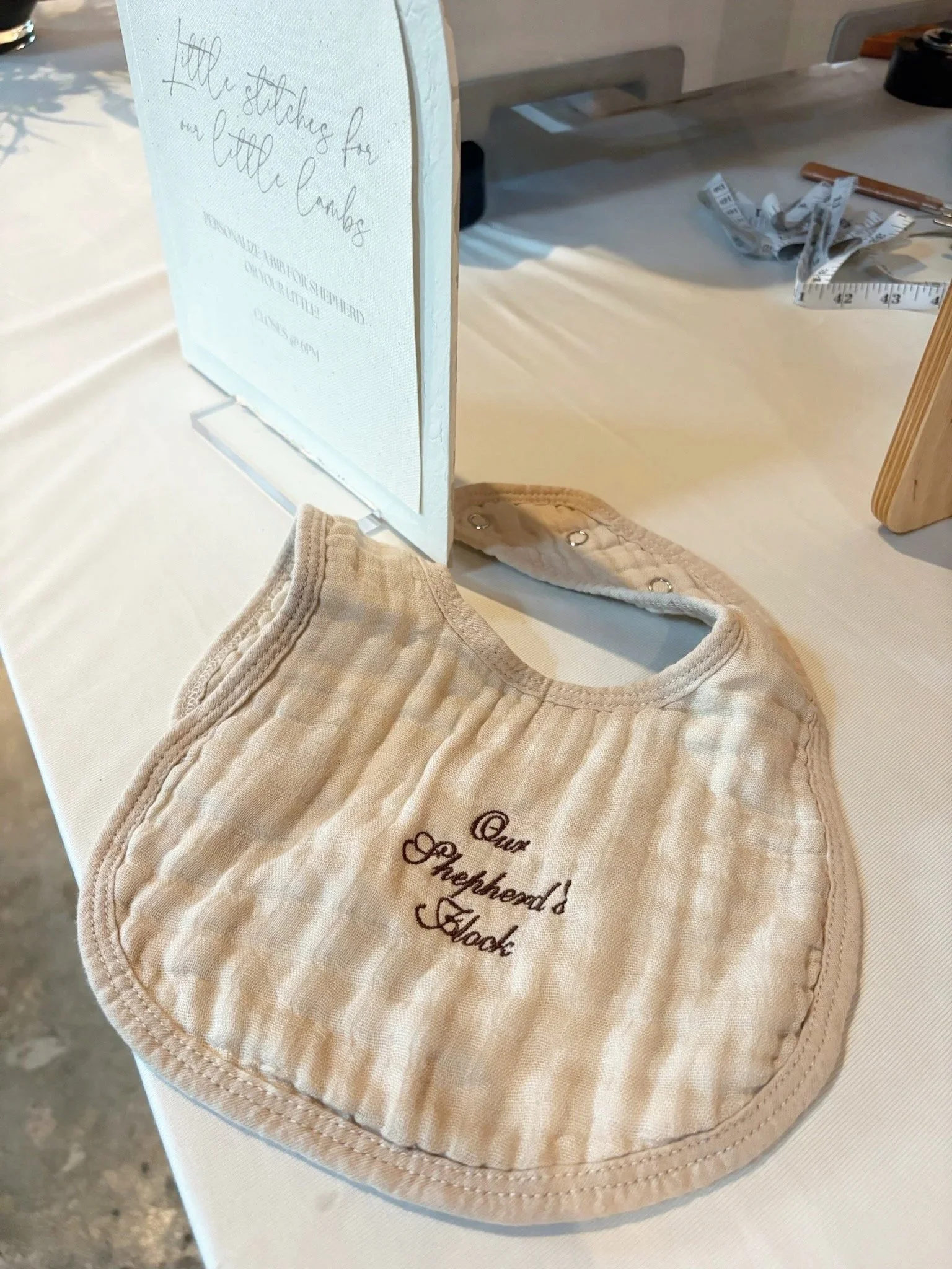 A beige quilted baby bib with embroidered text placed on a white table with sewing and measurement tools in the background.