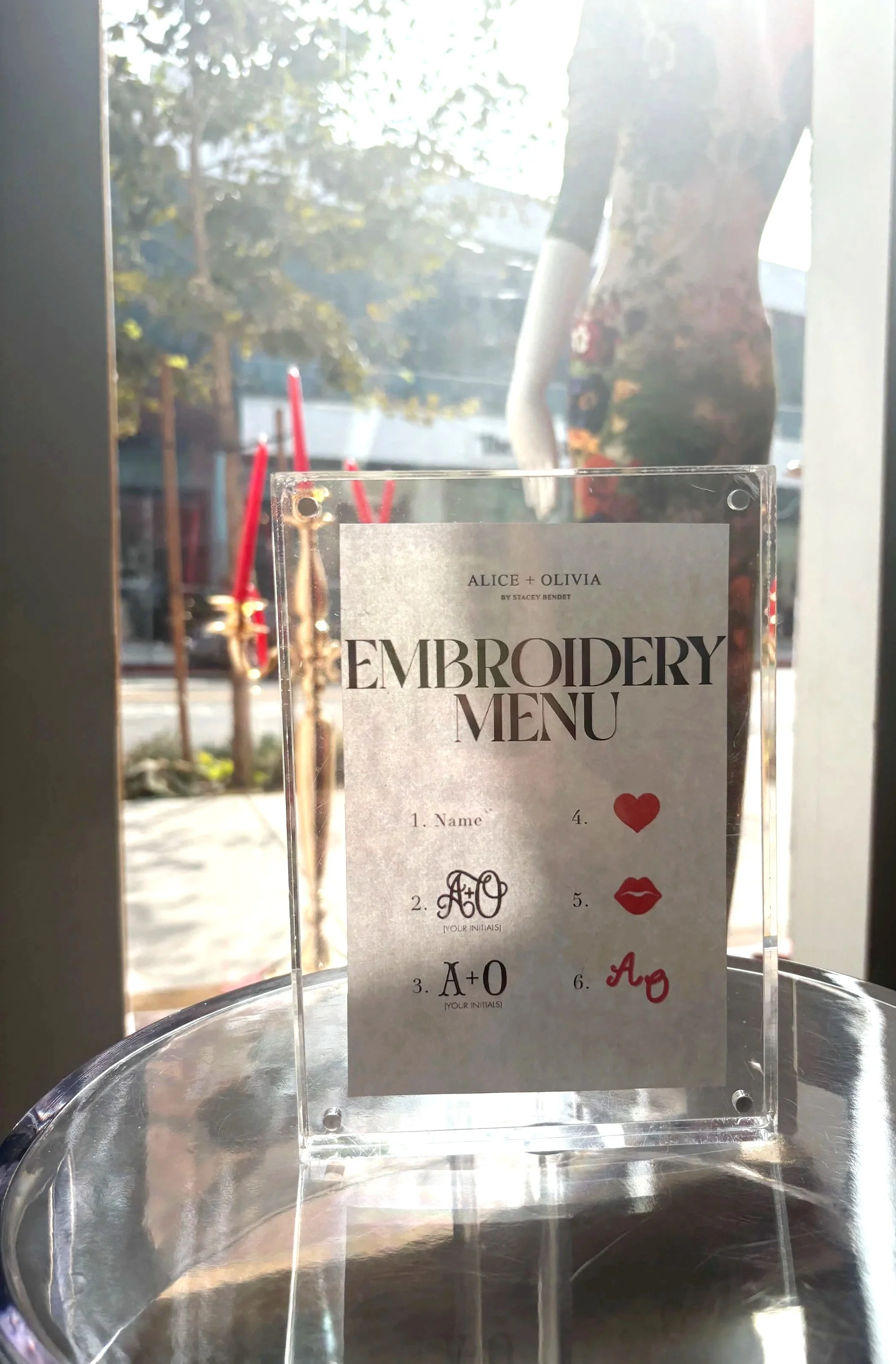 A clear acrylic embroidery menu stand displaying a menu with artistic text and symbols, placed on a reflective glass surface near a window with sunlight, tree, and outdoor scene visible in the background.