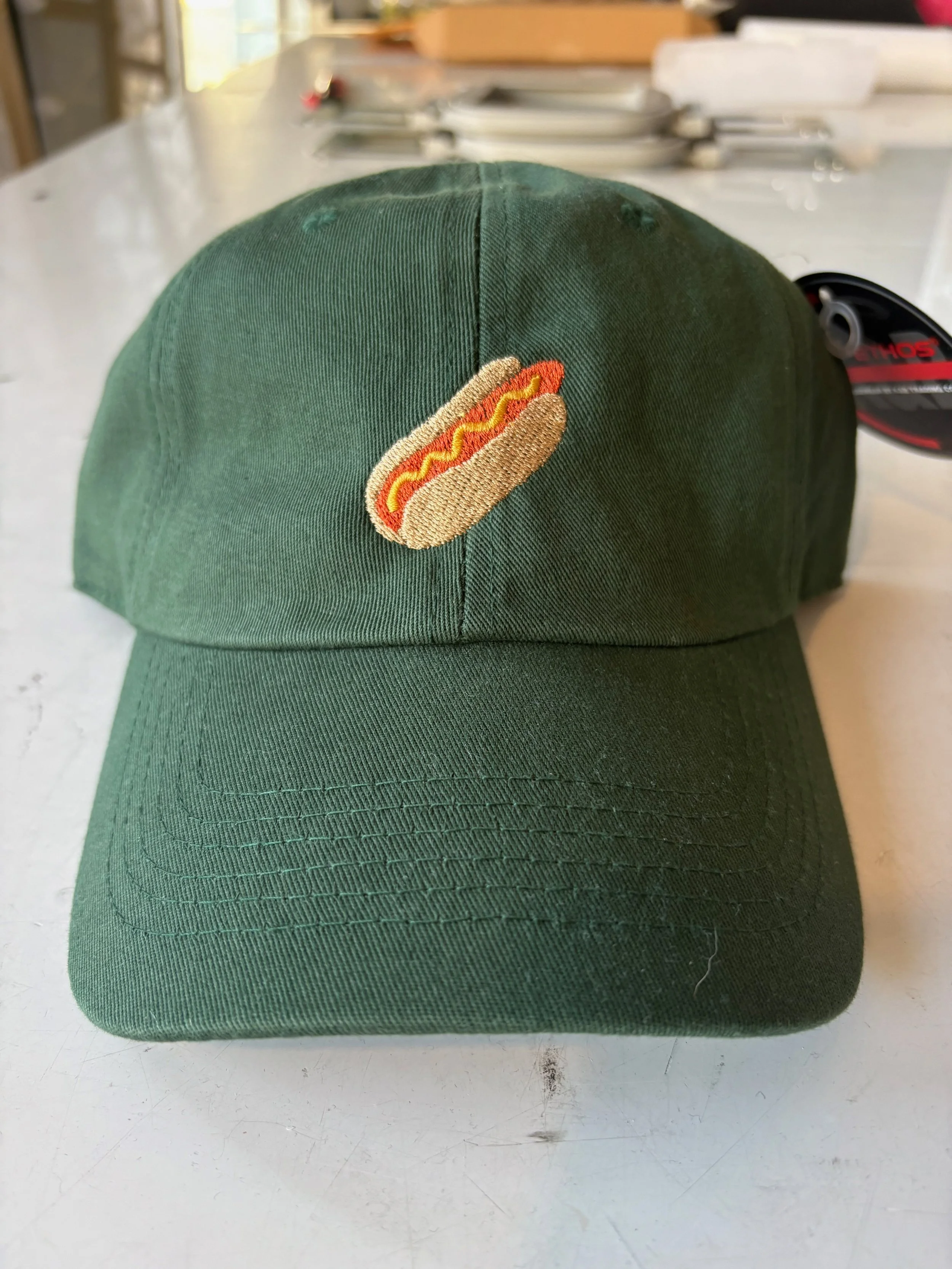 Green baseball cap with embroidered hot dog patch on front, placed on a white surface with utensils and papers in the background.