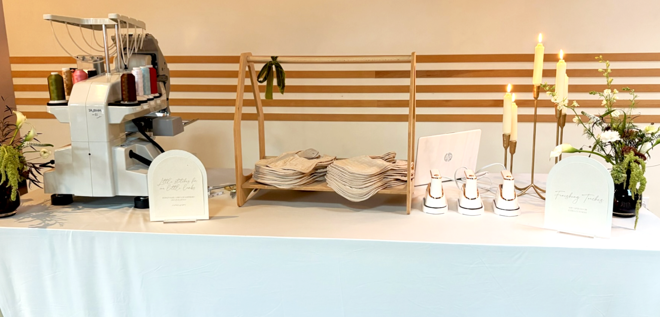 Event set-up for a luxury baby shower. A table with an embroidery machine, fabric stacks, candles, and floral arrangements.