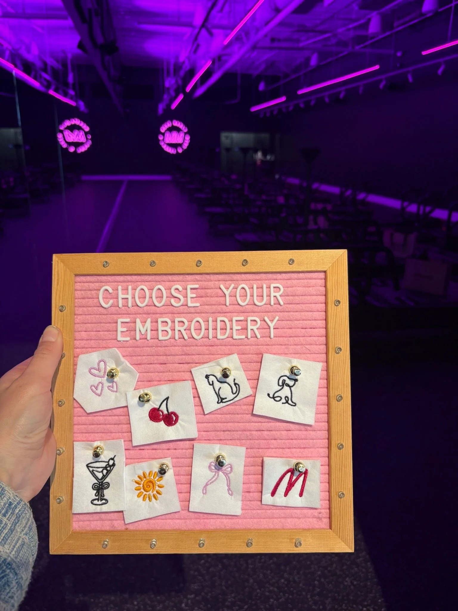 A person holding a pink letter board with embroidered images of cherries, a cocktail, a unicorn, a dolphin, a sun, a bow, and a red letter M, in a pilates studio.