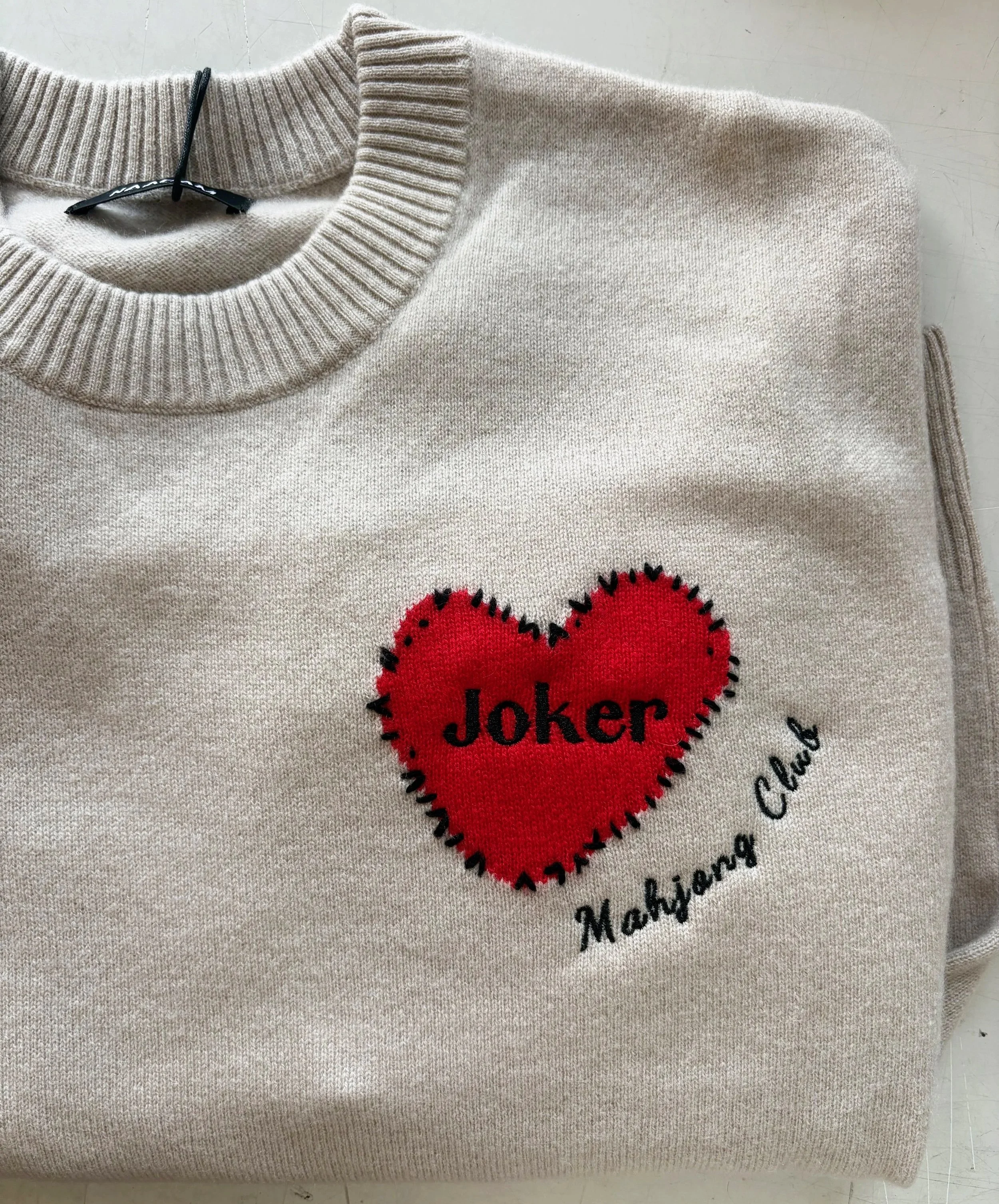 Close-up of a beige sweater with a red heart and black embroidered text that reads 'joker' on it, lying on a white surface.
