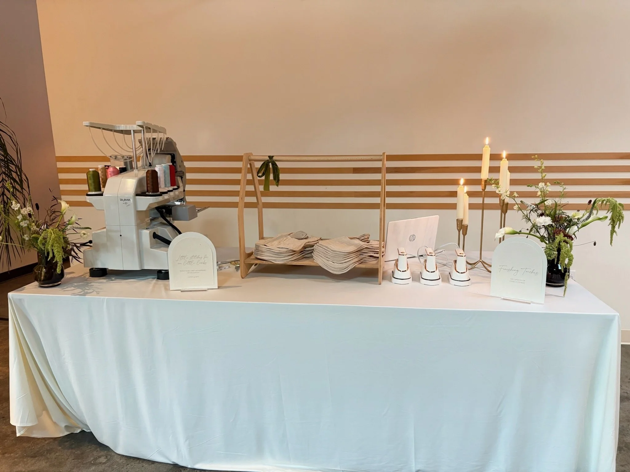 A table covered with a white tablecloth displays various floral arrangements, an embroidery machine, stacks of cloth, and lit candles.